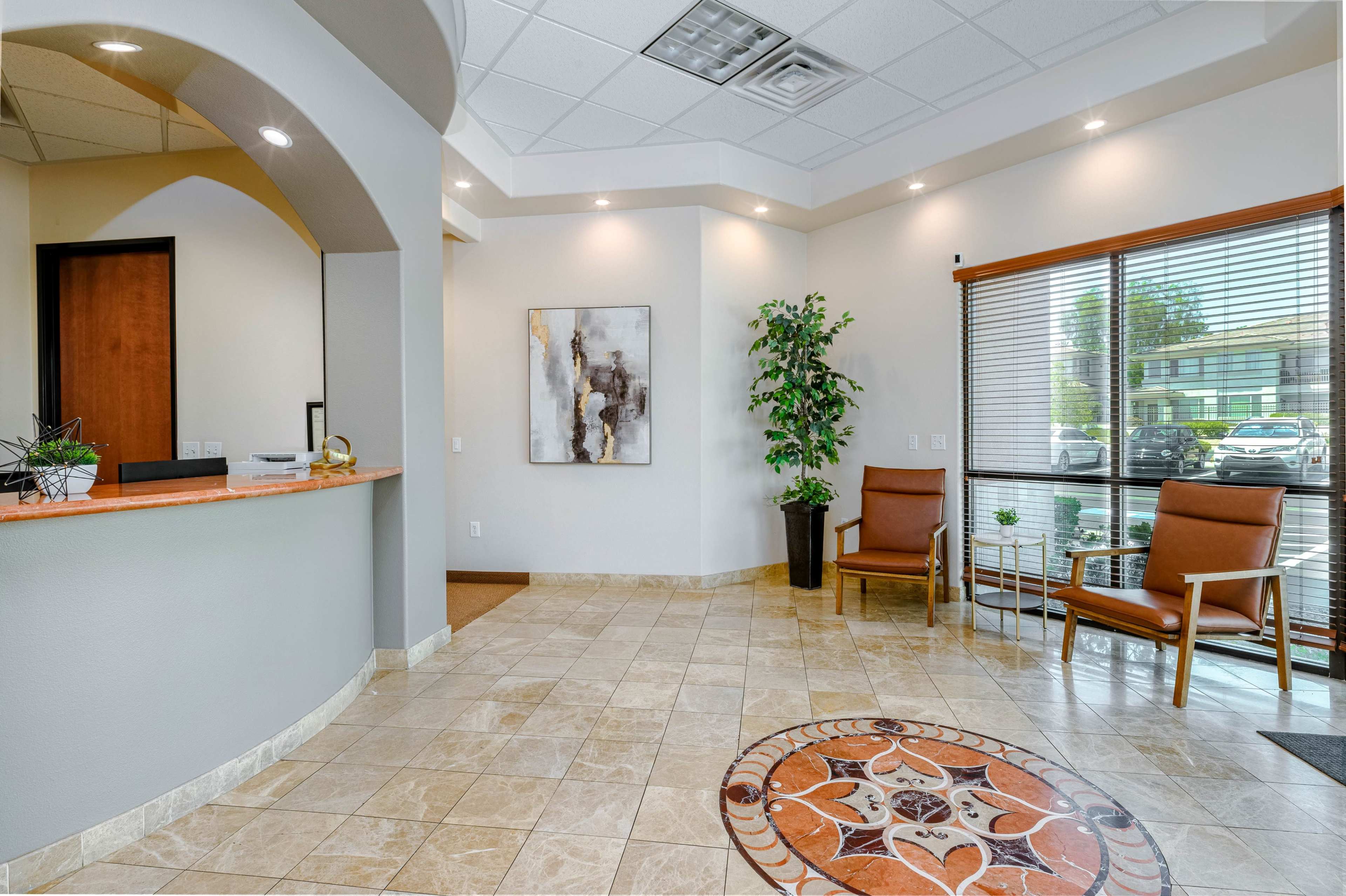 A modern reception area with a curved front desk, tiled flooring, and two chairs beside a potted plant and large windows.