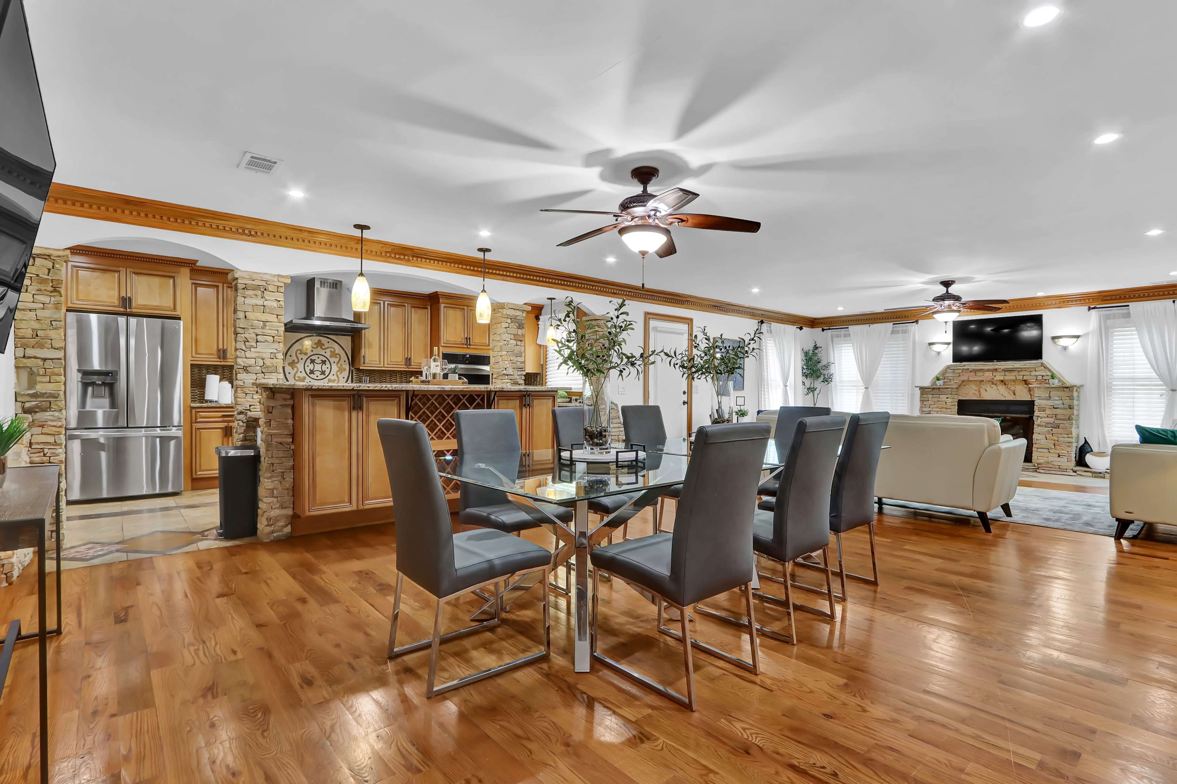 A modern, open-concept dining and living area featuring a wooden dining table with gray chairs, a stone fireplace, and a kitchen bar in the background.