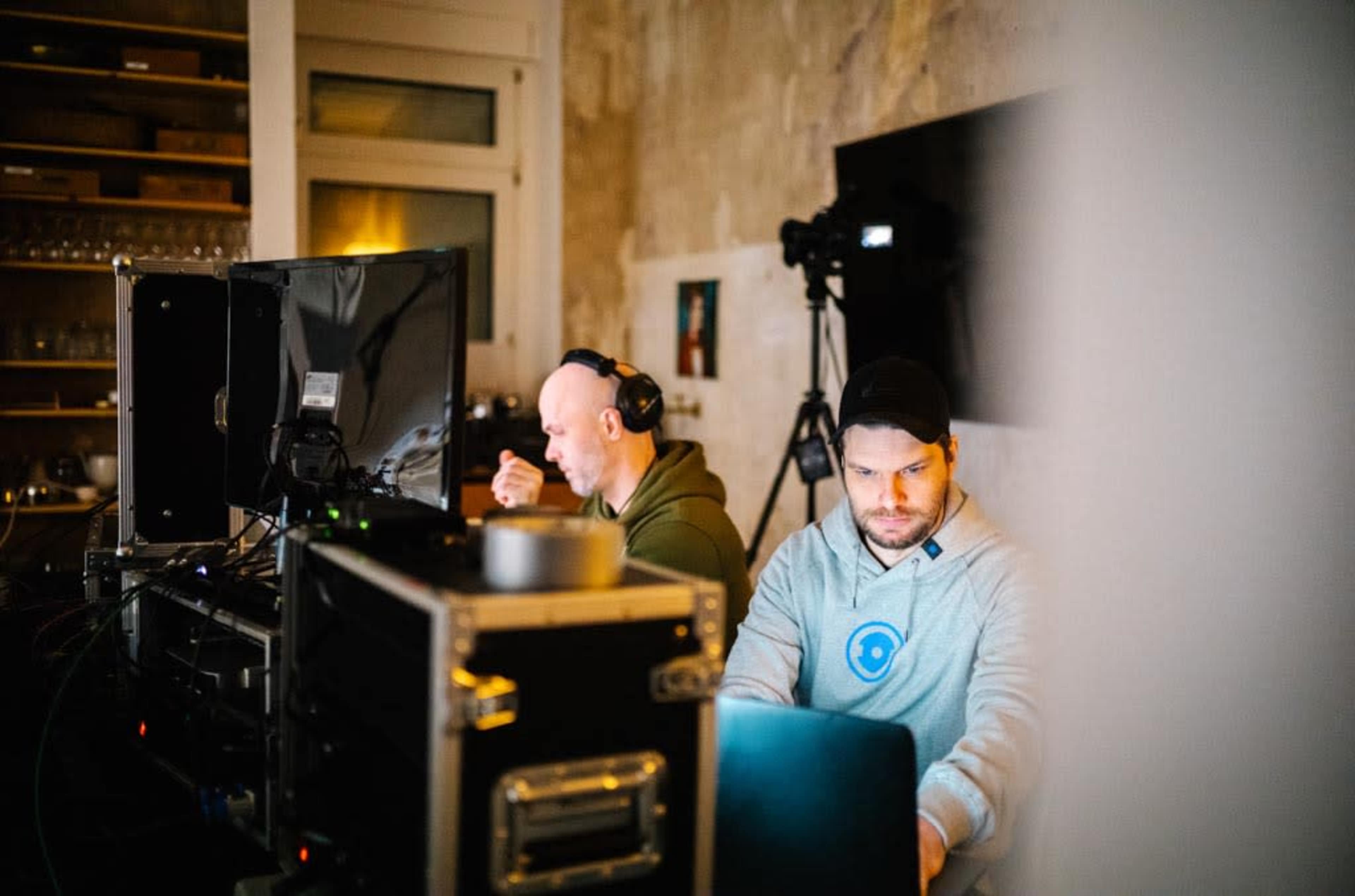 Two men work at computers in a room equipped with video production equipment and a camera.