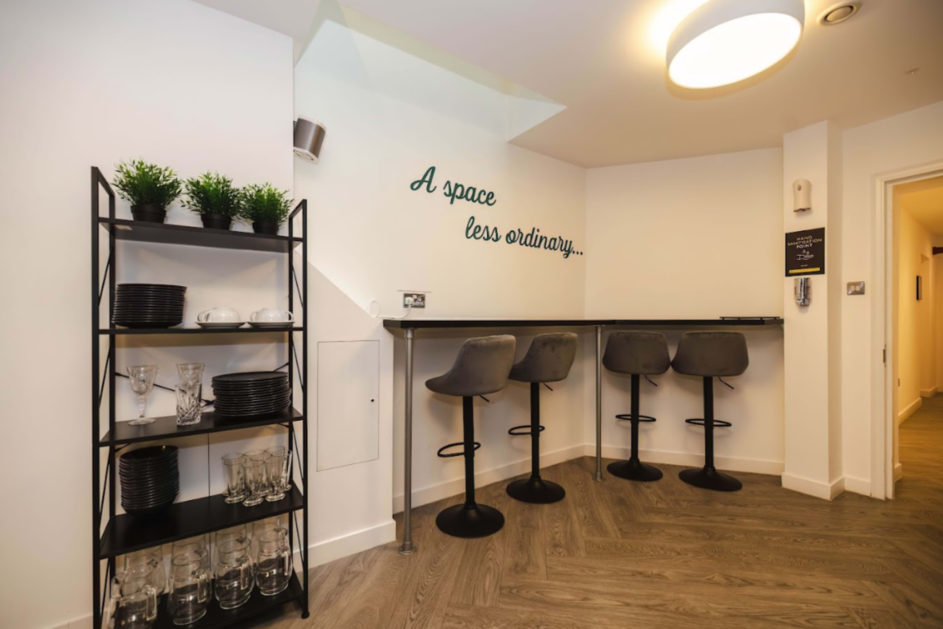 A modern kitchenette area with a black shelving unit filled with glassware and plates, alongside a bar counter with three stools and a wall featuring the phrase "A space less ordinary."