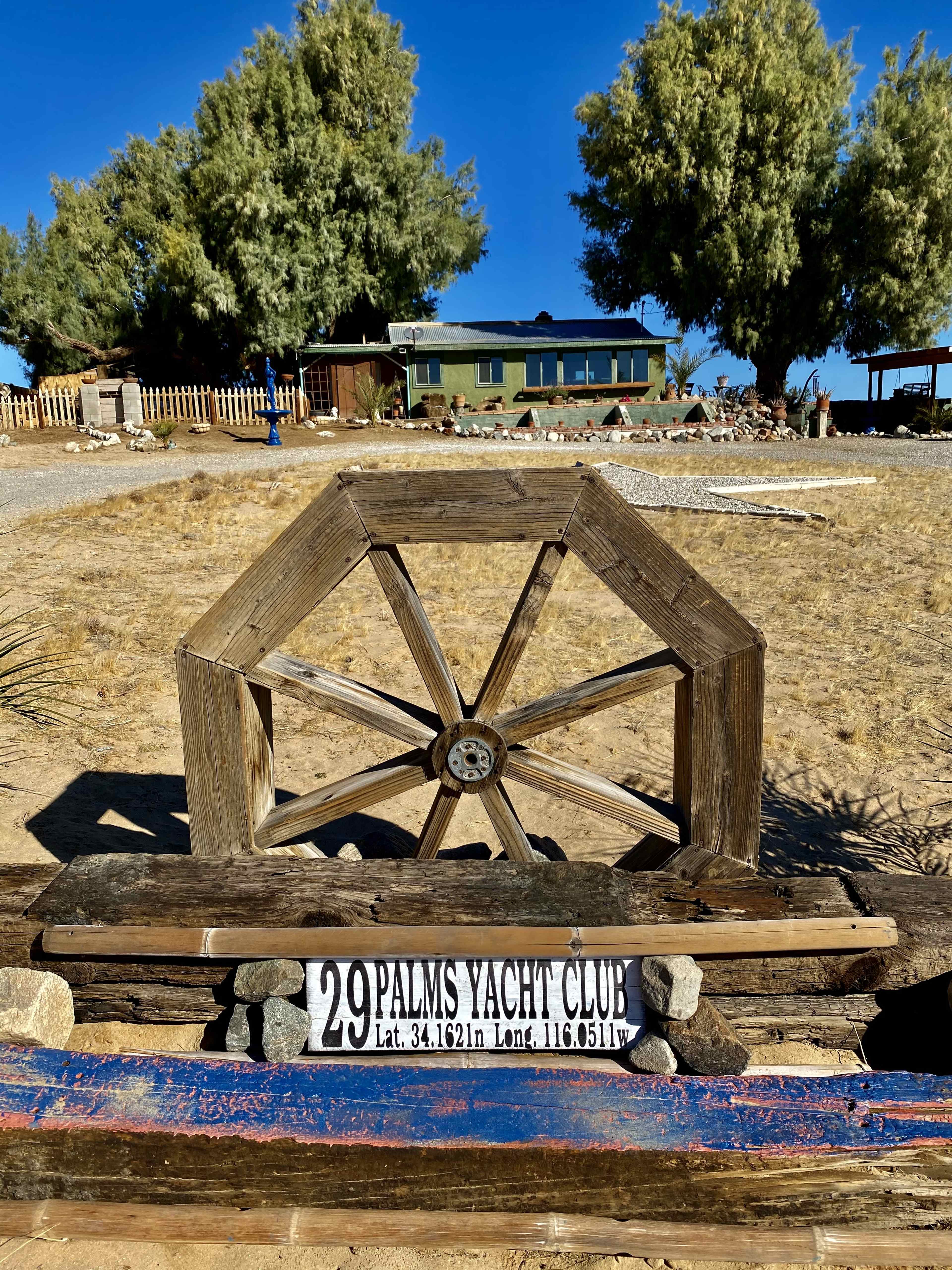 A wooden wheel structure is positioned in front of a sign for the 29 Palms Yacht Club, with a house and trees visible in the background.