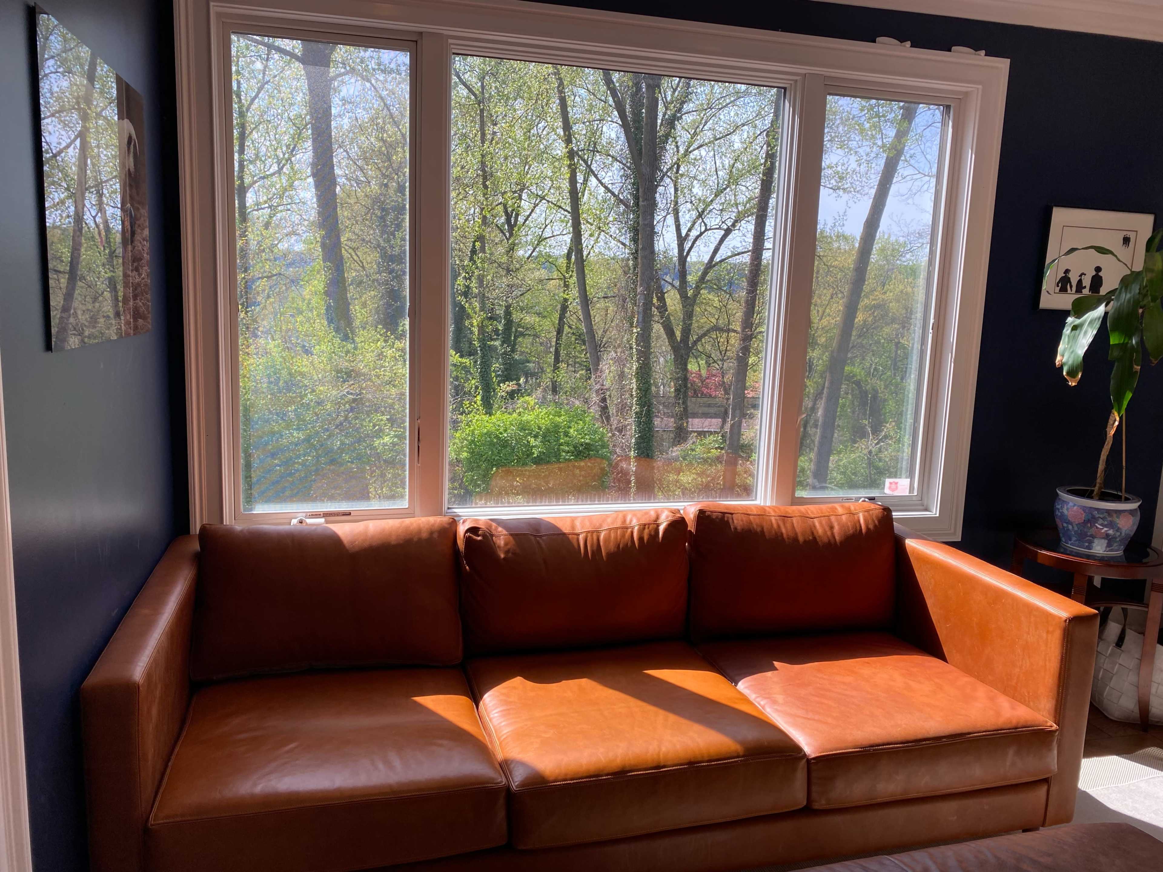 A brown leather couch is positioned in front of large windows overlooking a wooded area.