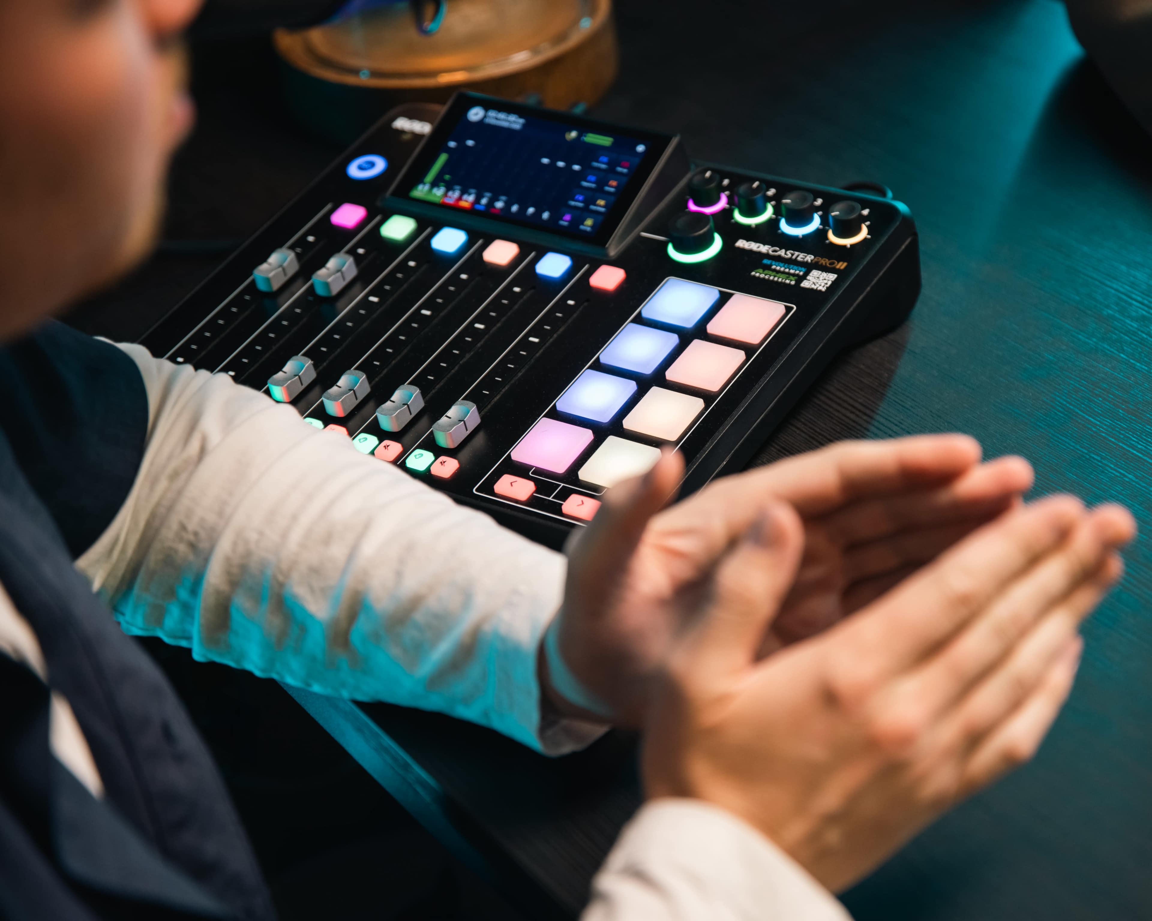 A person is seen with their hands clapped together next to a digital audio mixer featuring colorful buttons and sliders.