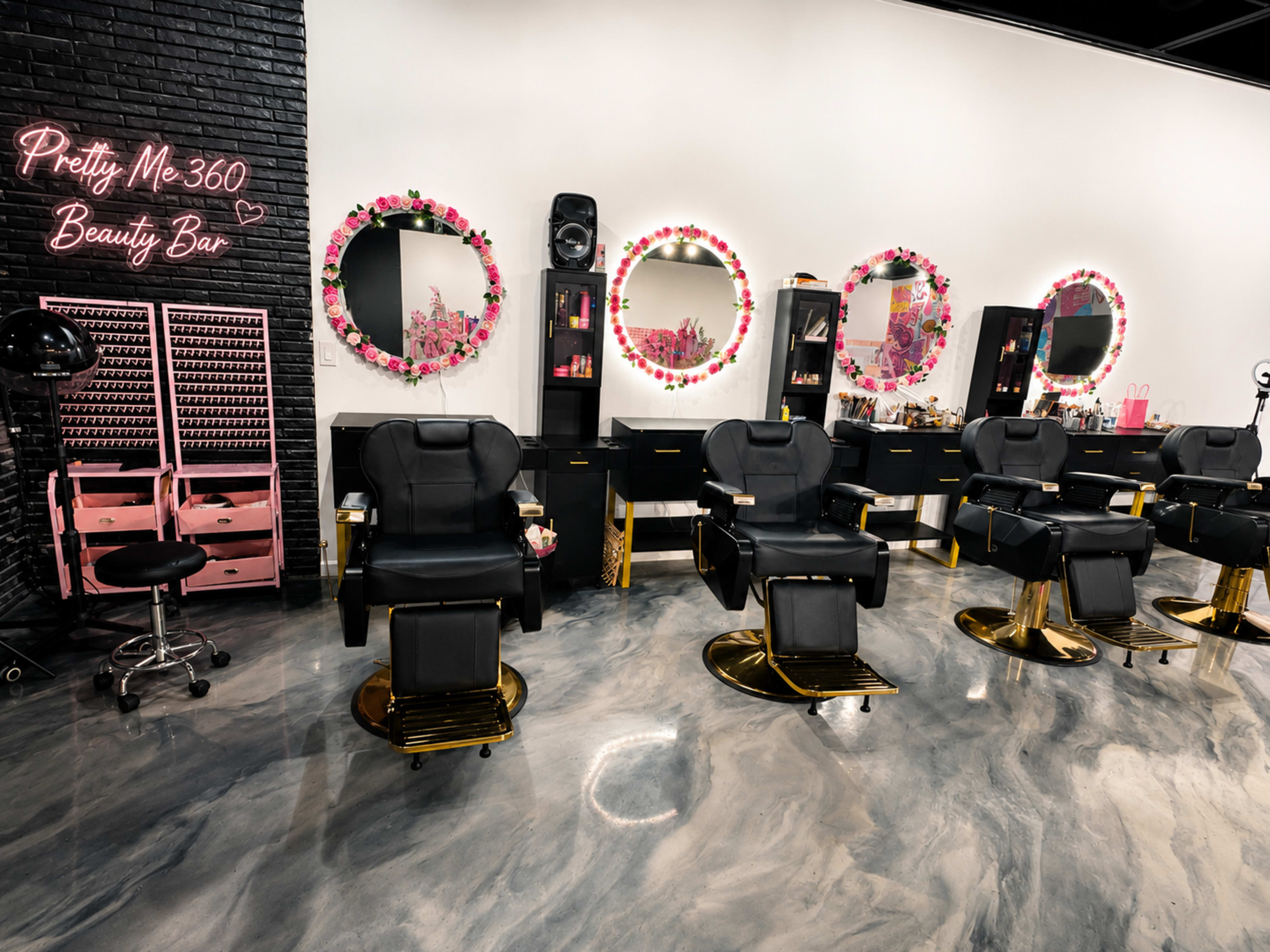 The image shows a modern beauty bar with black and gold styling chairs, illuminated mirrors, and a decorative wall featuring the name "Pretty Me 360 Beauty Bar."