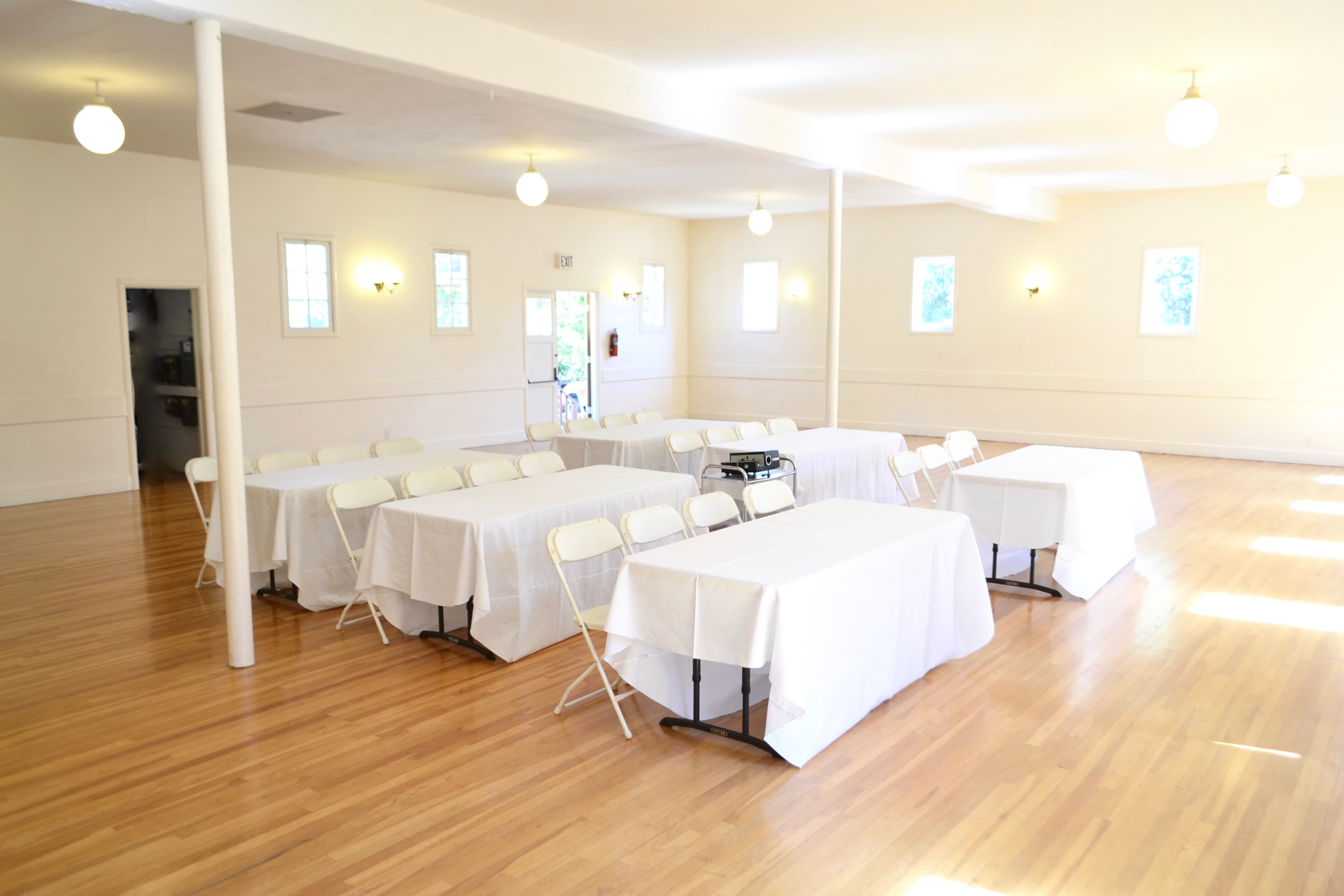 A spacious room with wooden floors arranged with multiple tables covered in white cloth and surrounded by folding chairs.