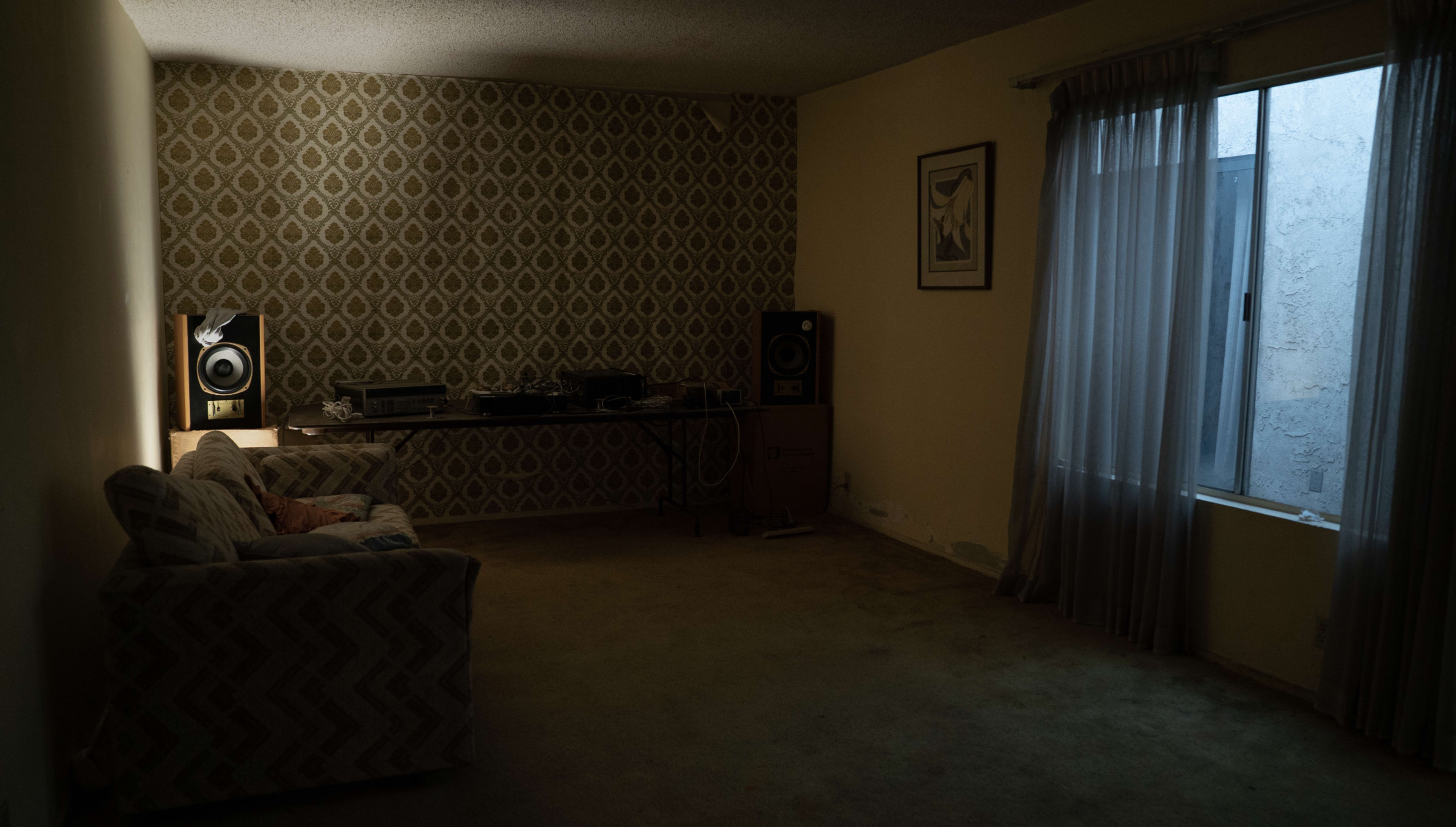 An empty living room features a patterned wall, a sofa, a table with audio equipment, and large curtains over a window.