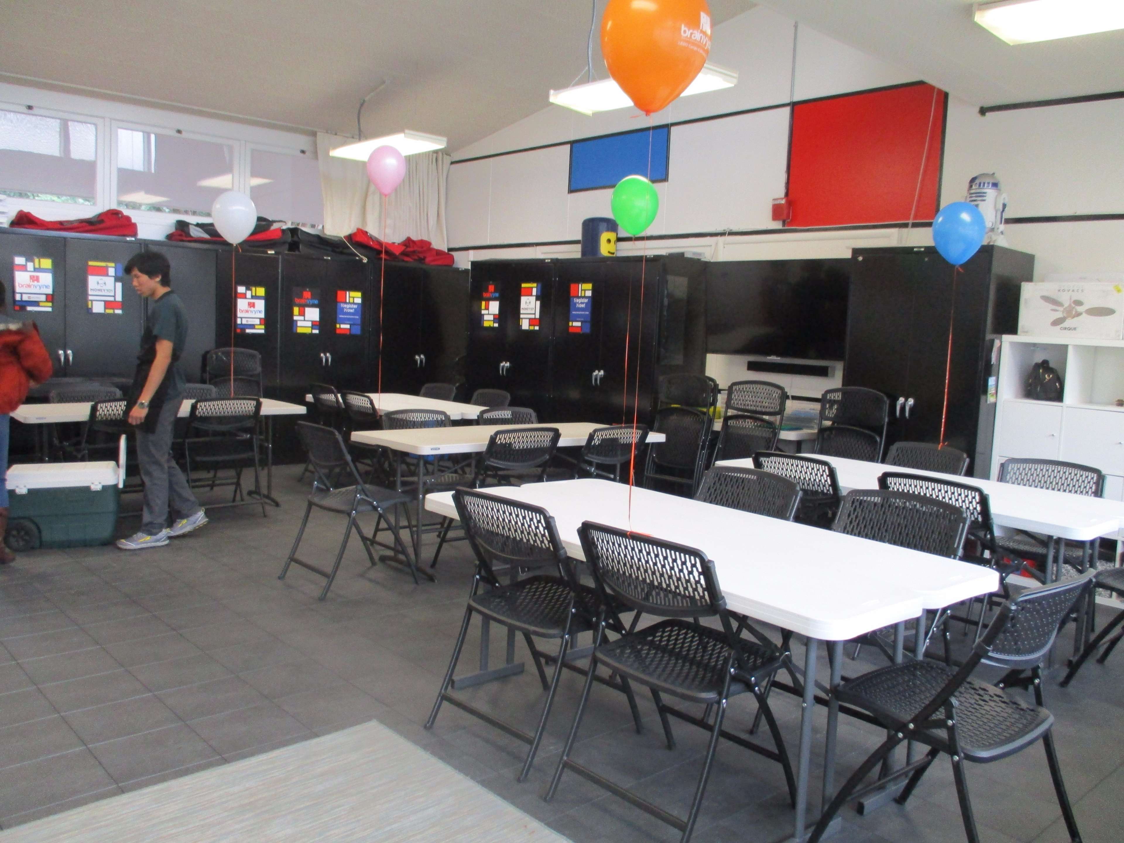 A spacious room with several tables and chairs, decorated with balloons, and featuring black cabinets along the walls.