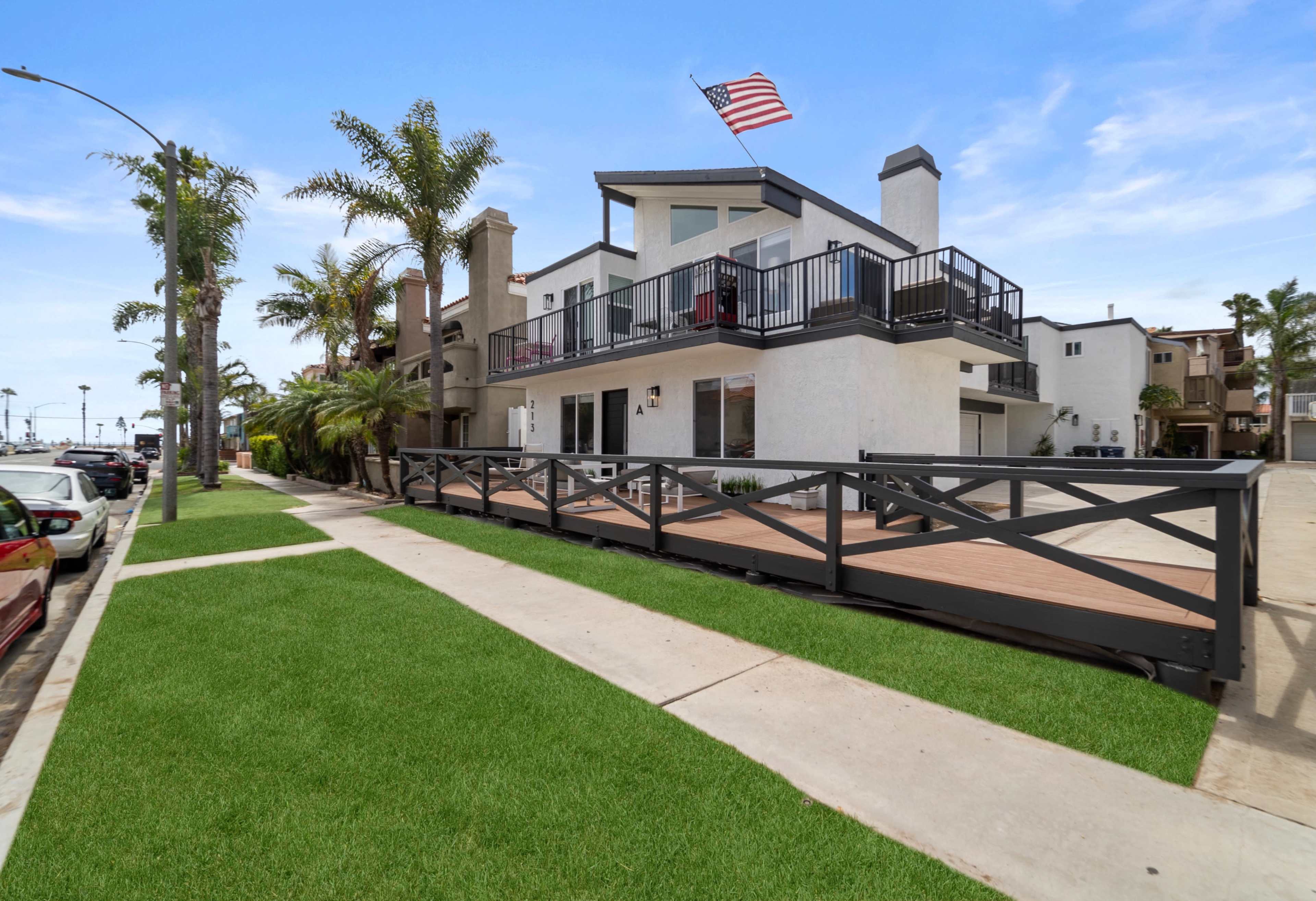 A modern two-story building with a balcony, surrounded by palm trees, and featuring a front patio deck, is located along a paved sidewalk.