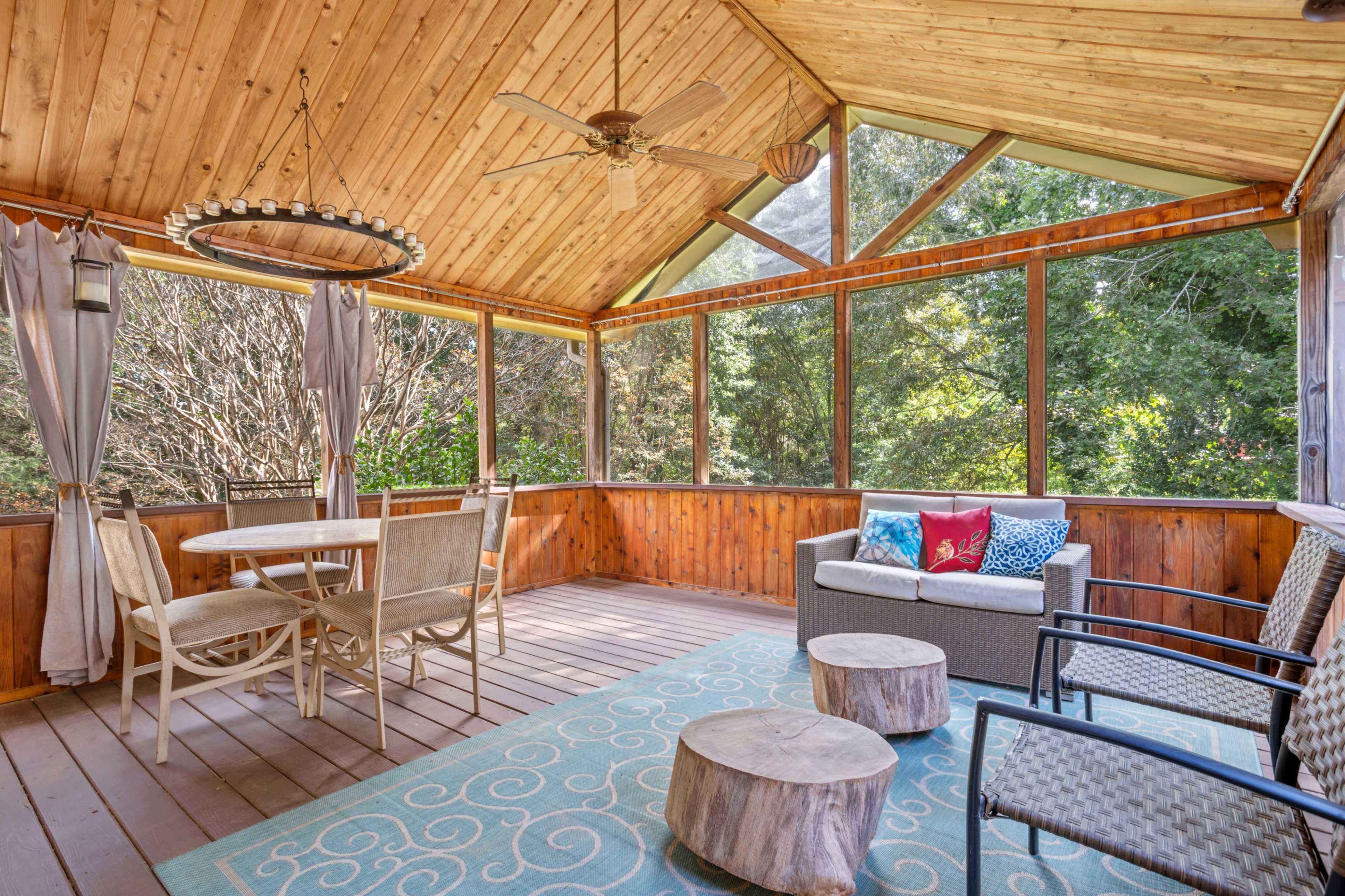 A screened-in porch with wooden walls and ceiling, featuring a dining table, seating area, and a patterned rug.