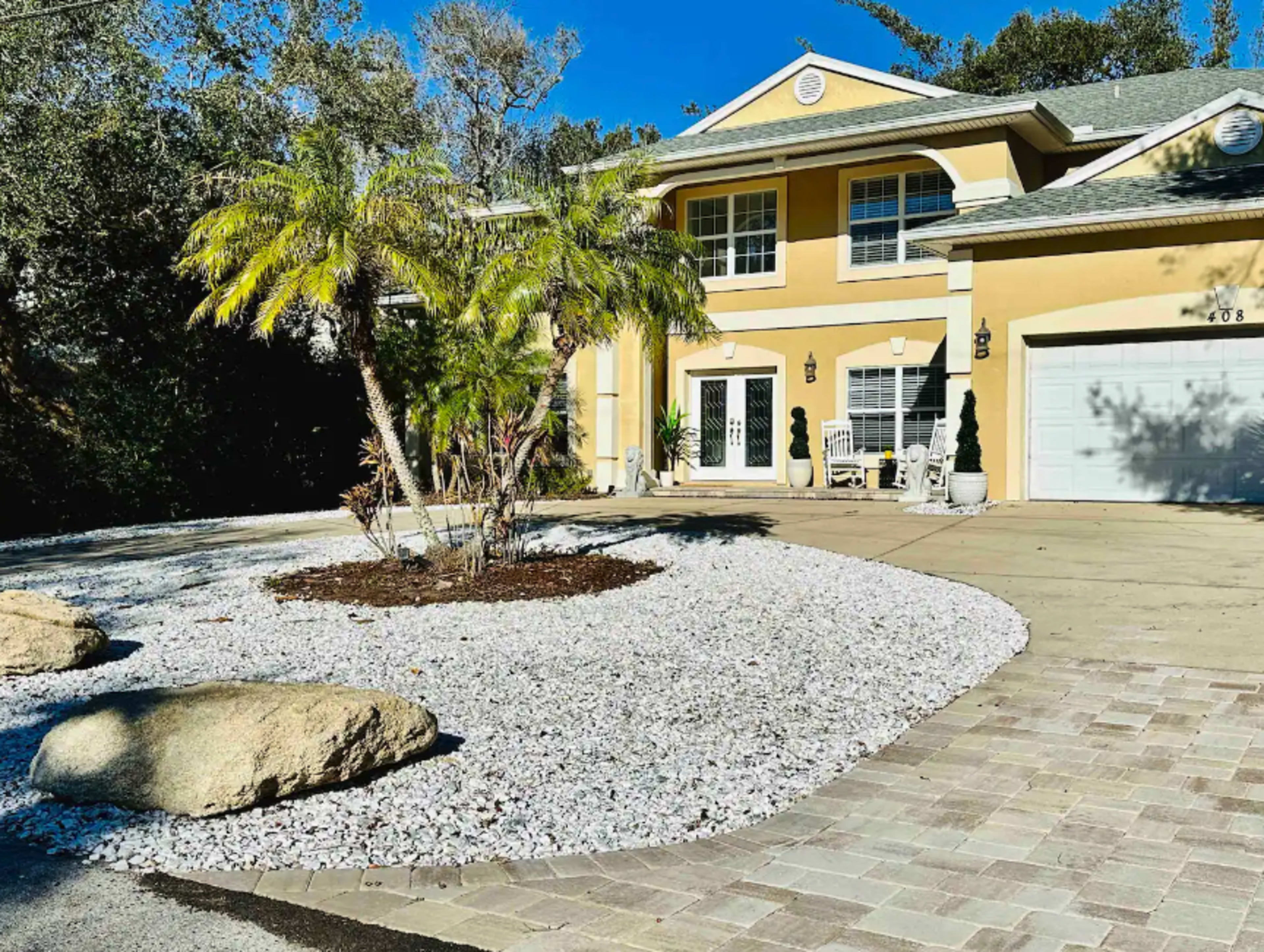 A two-story yellow house with a landscaped front yard featuring decorative rocks and a palm tree.