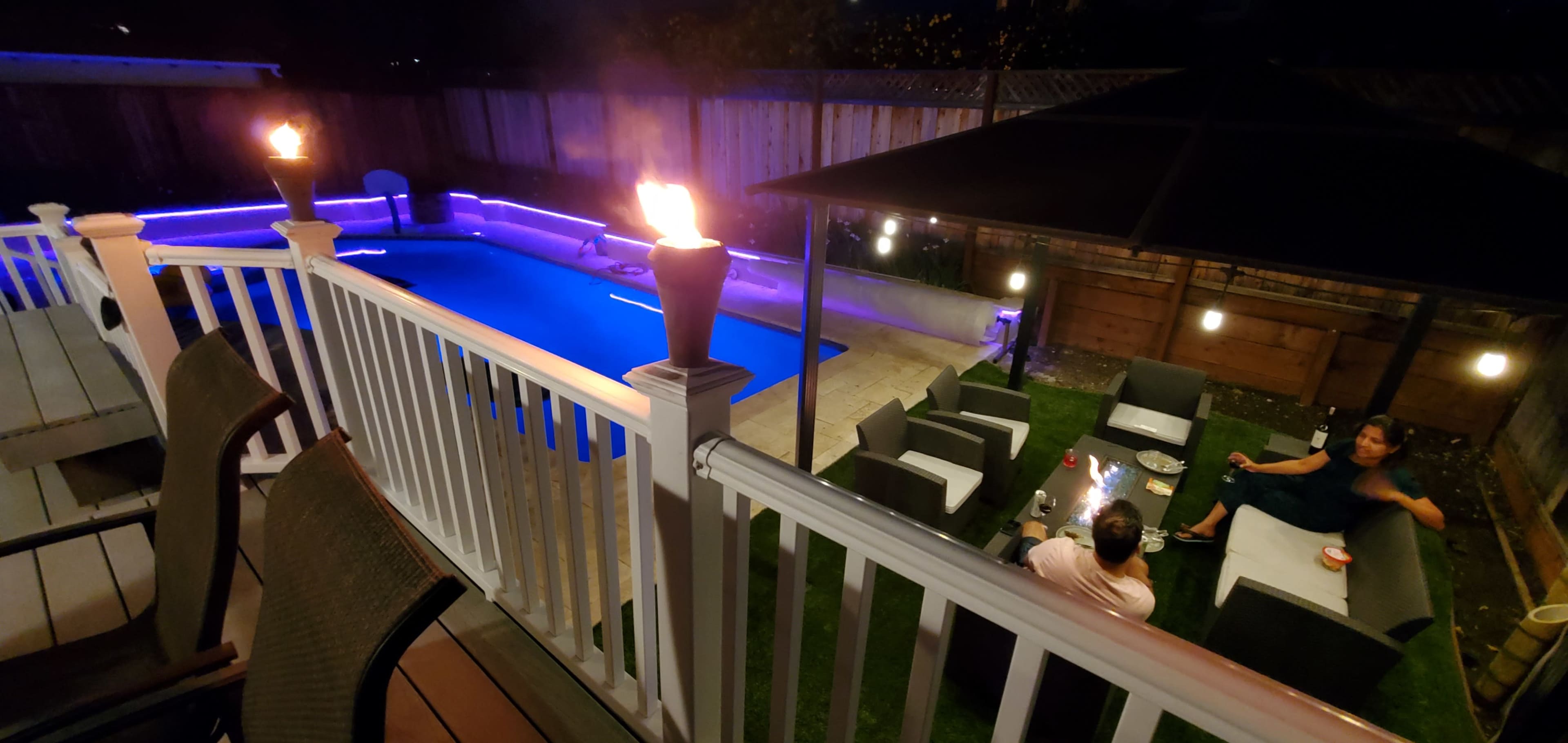 A backyard scene features a brightly lit pool surrounded by patio furniture, with torches illuminating the area and two people seated nearby.