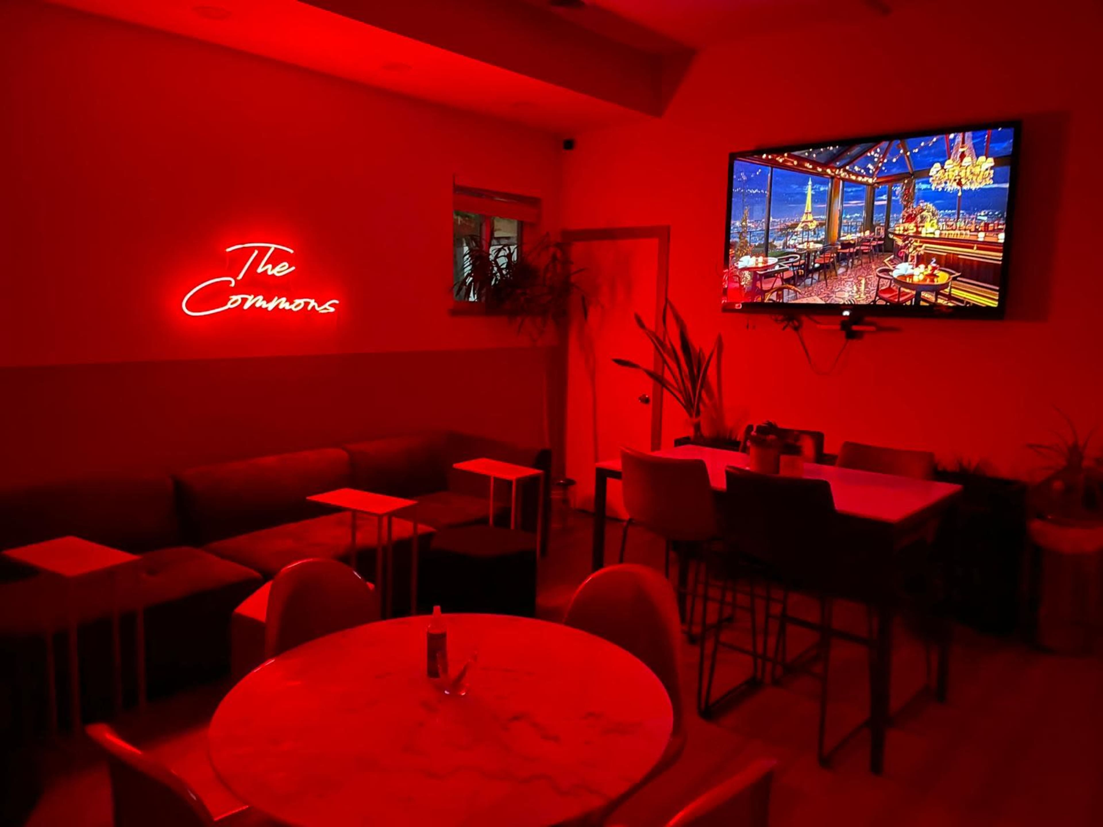 The interior of a lounge features red lighting, a neon sign that reads "The Connoisseurs," a large television displaying a scenic view, and various seating arrangements.