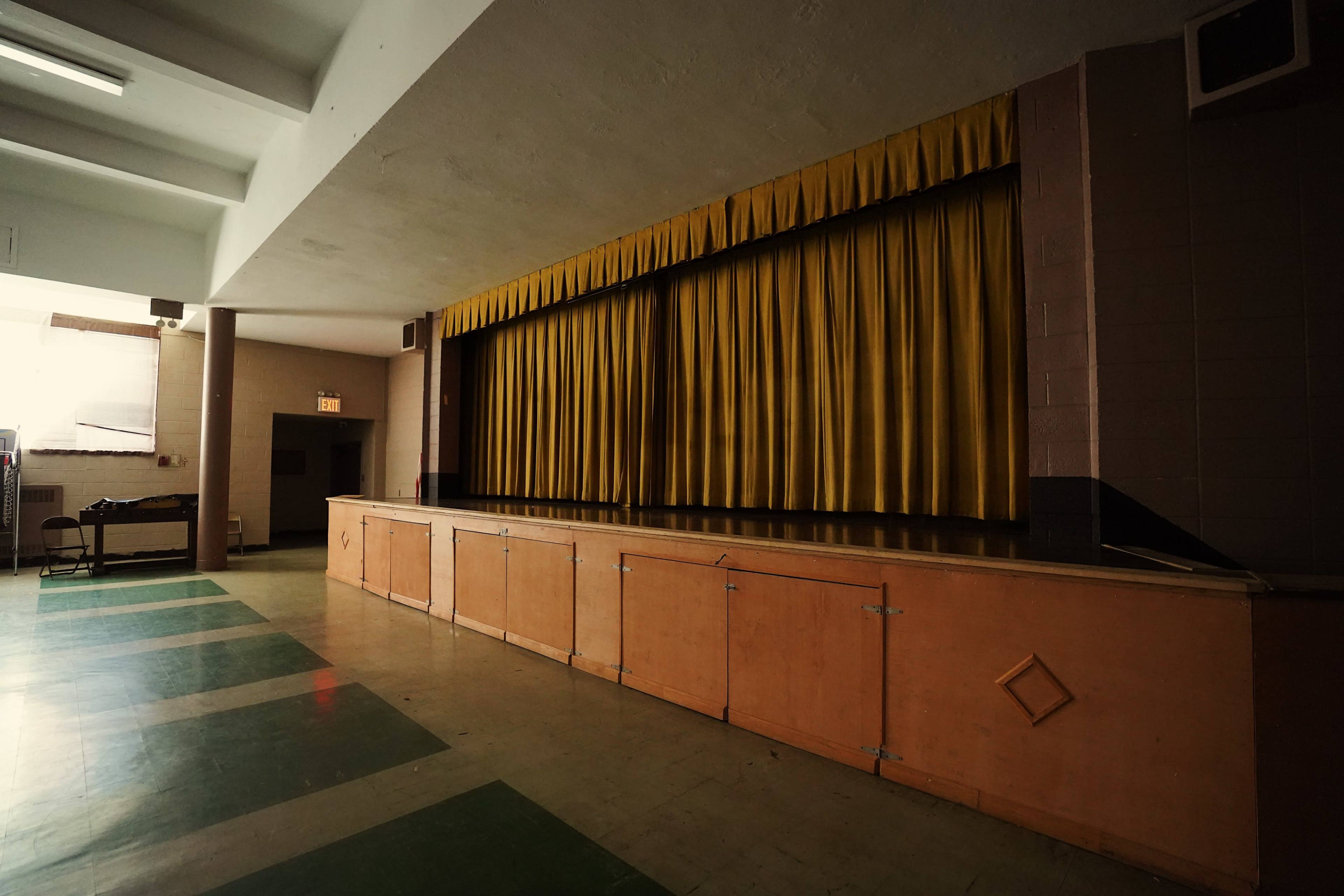 An empty stage with yellow curtains in a room featuring a wooden platform and green and gray-tiled flooring.
