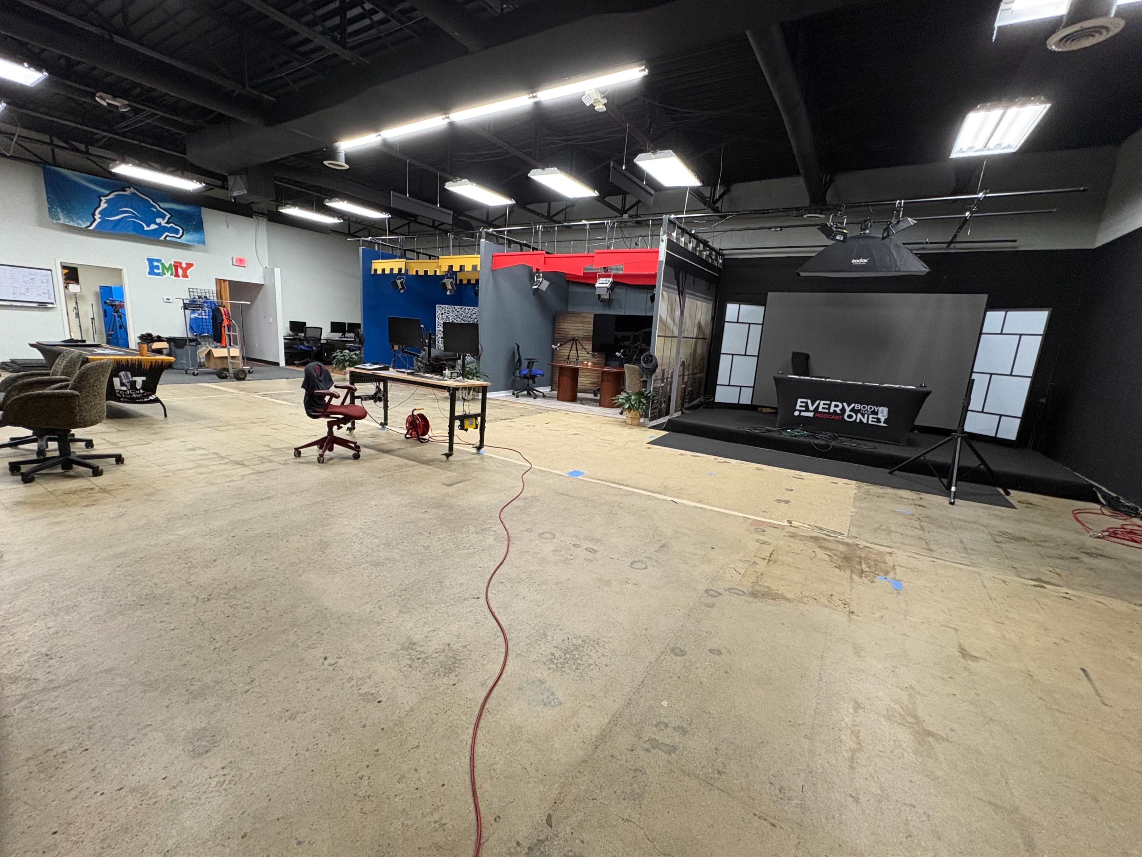 The image shows a spacious studio environment with a partially set-up broadcast area, scattered furniture, and a concrete floor.