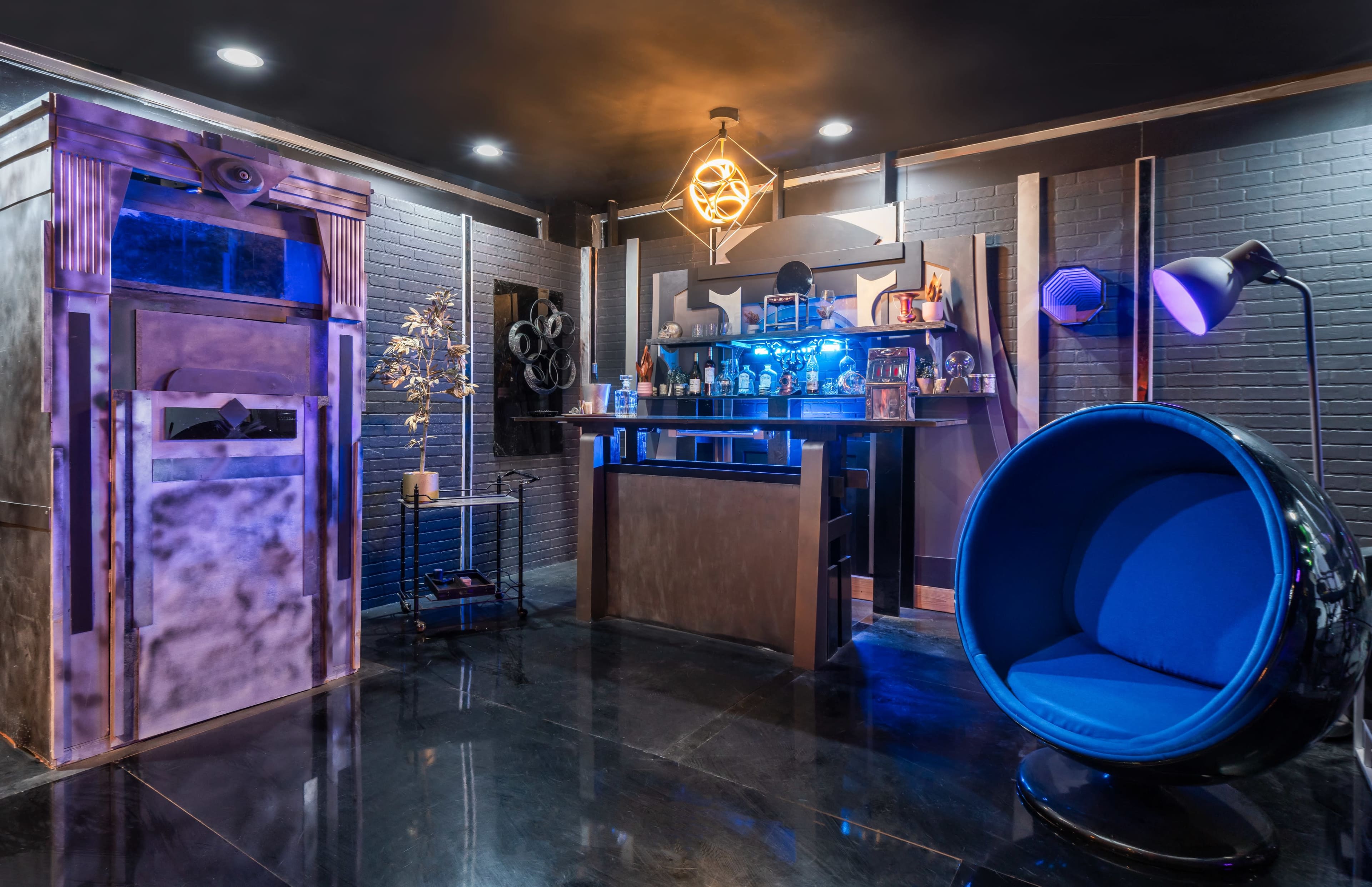 A modern bar area with a dark color scheme, featuring a curved blue chair, a sleek bar with illuminated shelves, and decorative elements on the walls.
