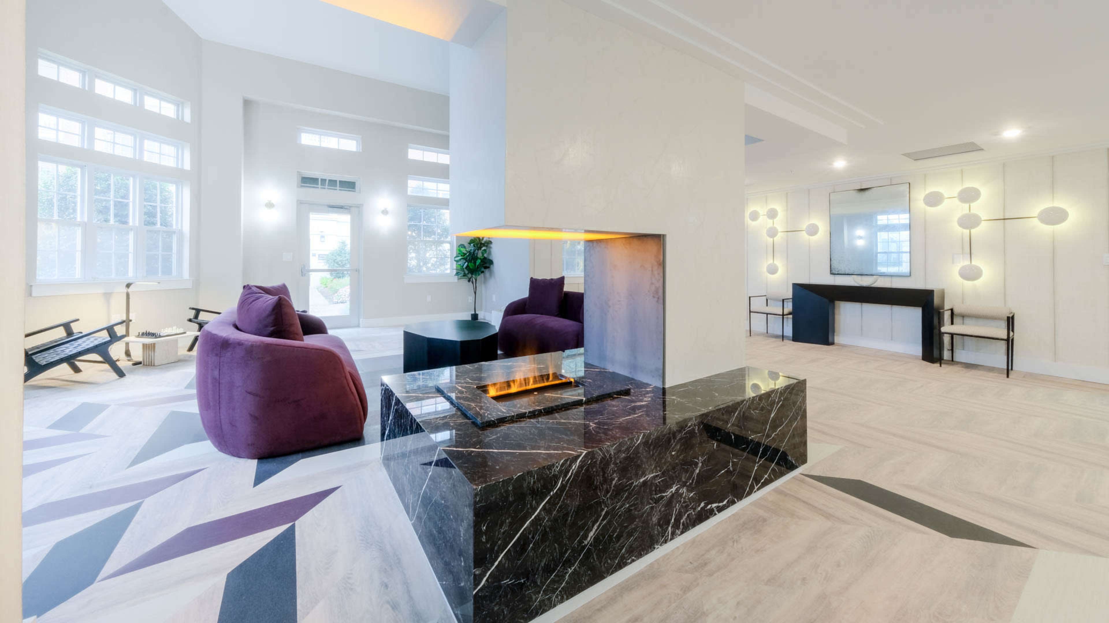 A modern lobby area featuring a central fireplace surrounded by purple armchairs, large windows, and a sleek, contemporary design.