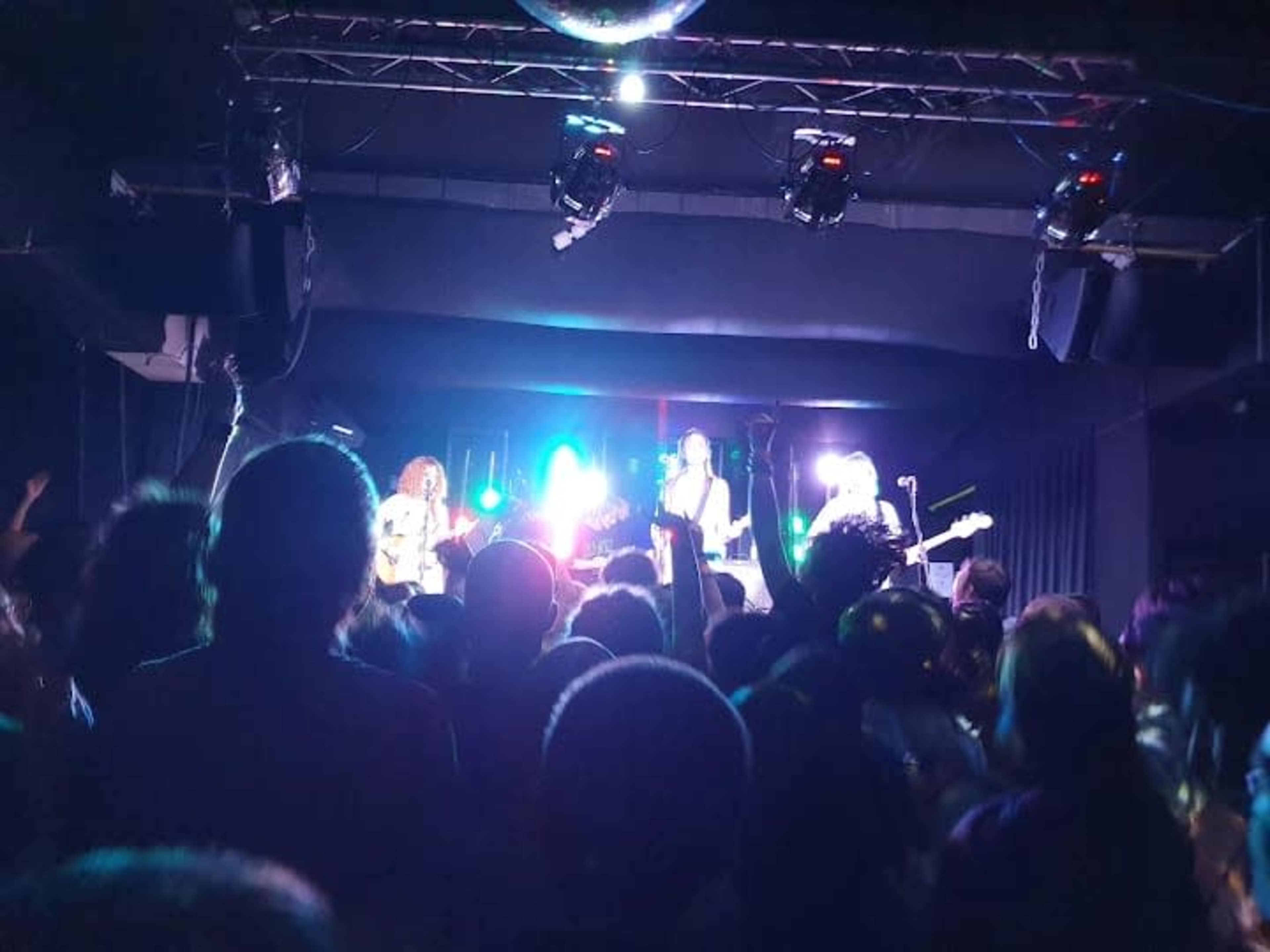 A band performs on stage in front of a lively audience with colorful lights illuminating the scene.