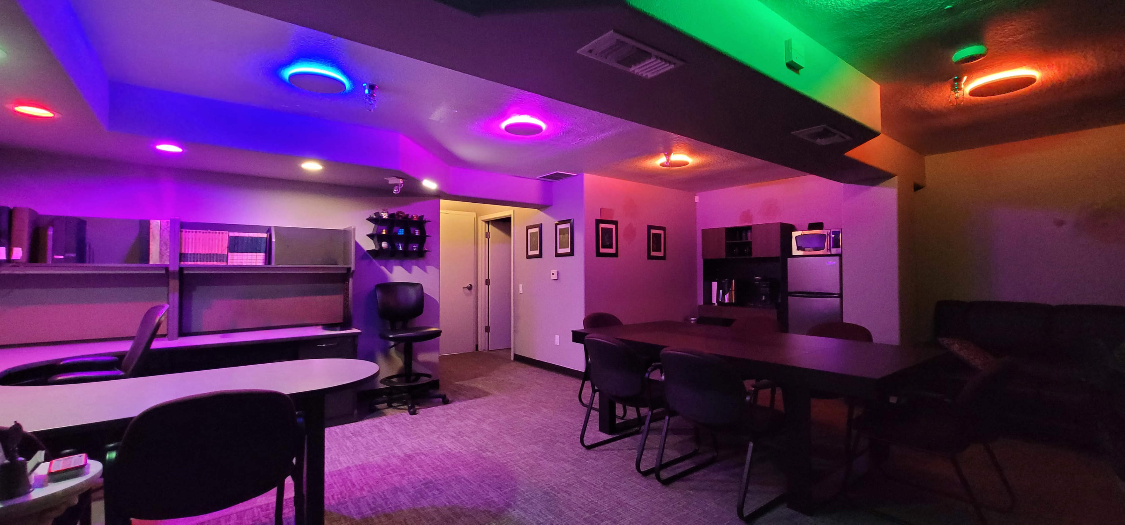 The image shows a dimly lit room with colorful circular LED lights on the ceiling, featuring a table surrounded by chairs, a desk, shelves, and various appliances.