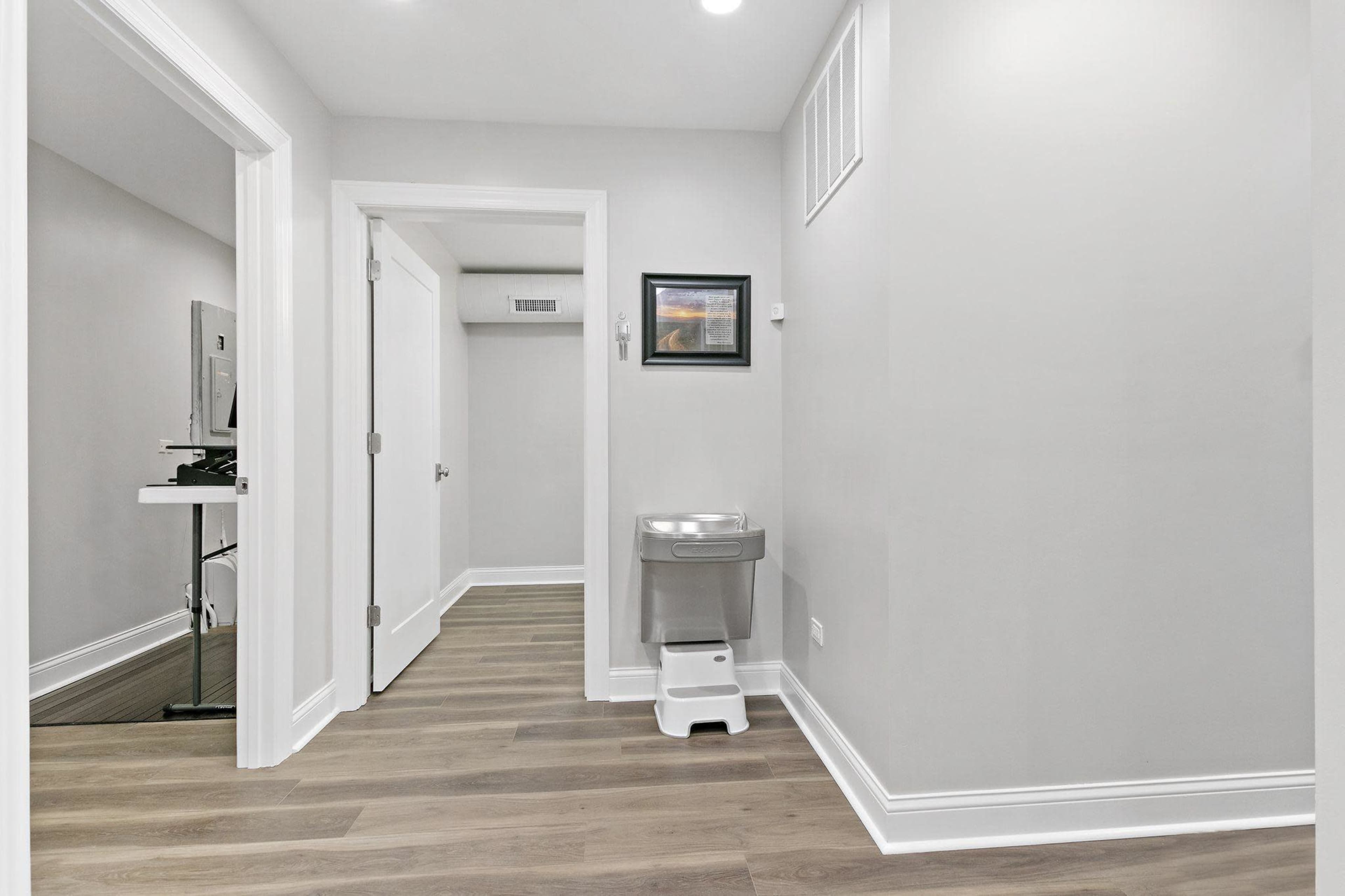 A modern hallway with light gray walls, vinyl flooring, and a water fountain next to a framed picture.