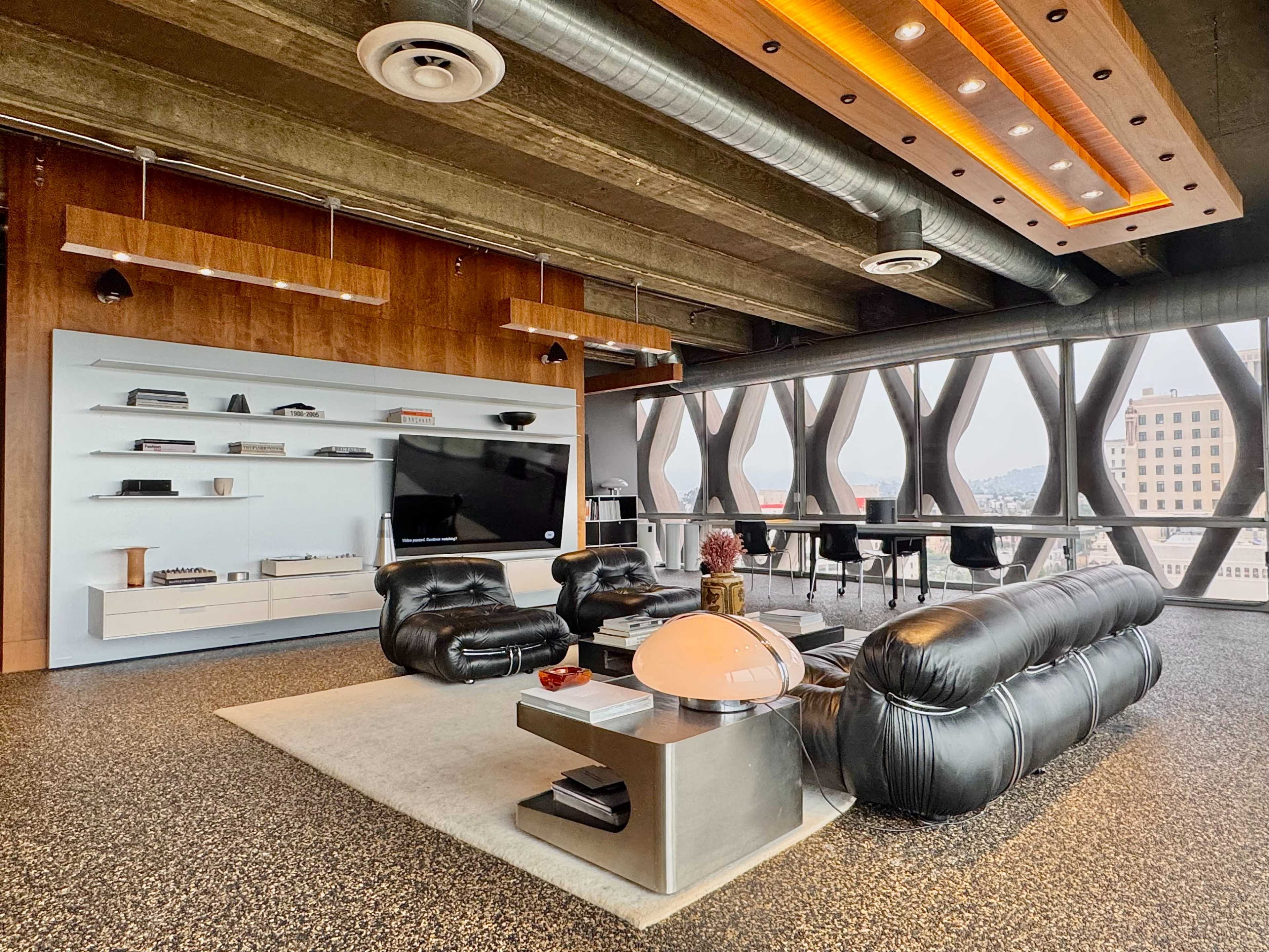 The image shows a modern living area with a sleek black sofa, a television mounted on a wall, and large windows revealing a city view.