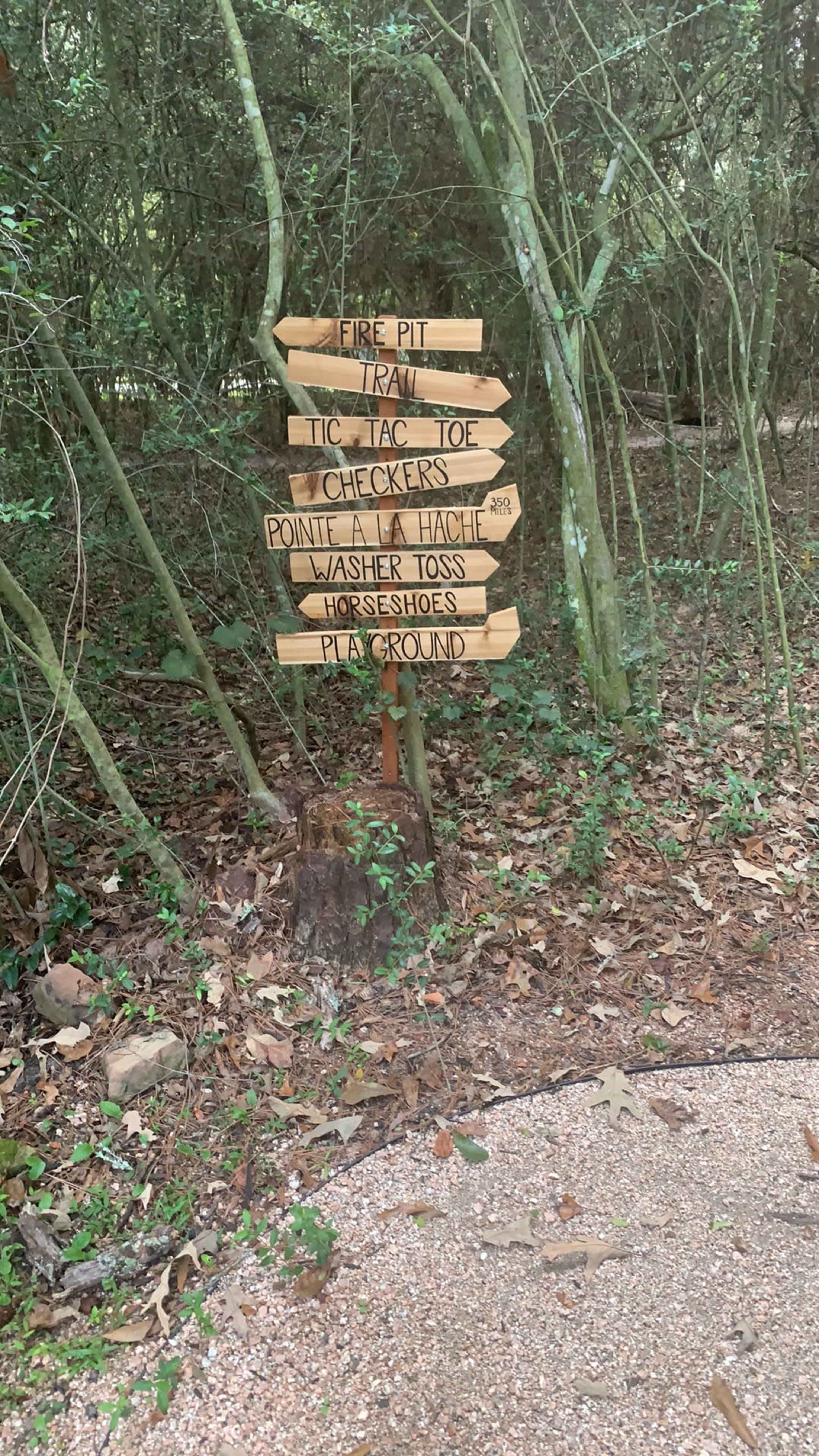 A wooden signpost lists various activities available at a park, including fire pit, train, tic tac toe, checkers, point a la hache, washer toss, horseshoes, and a playground.