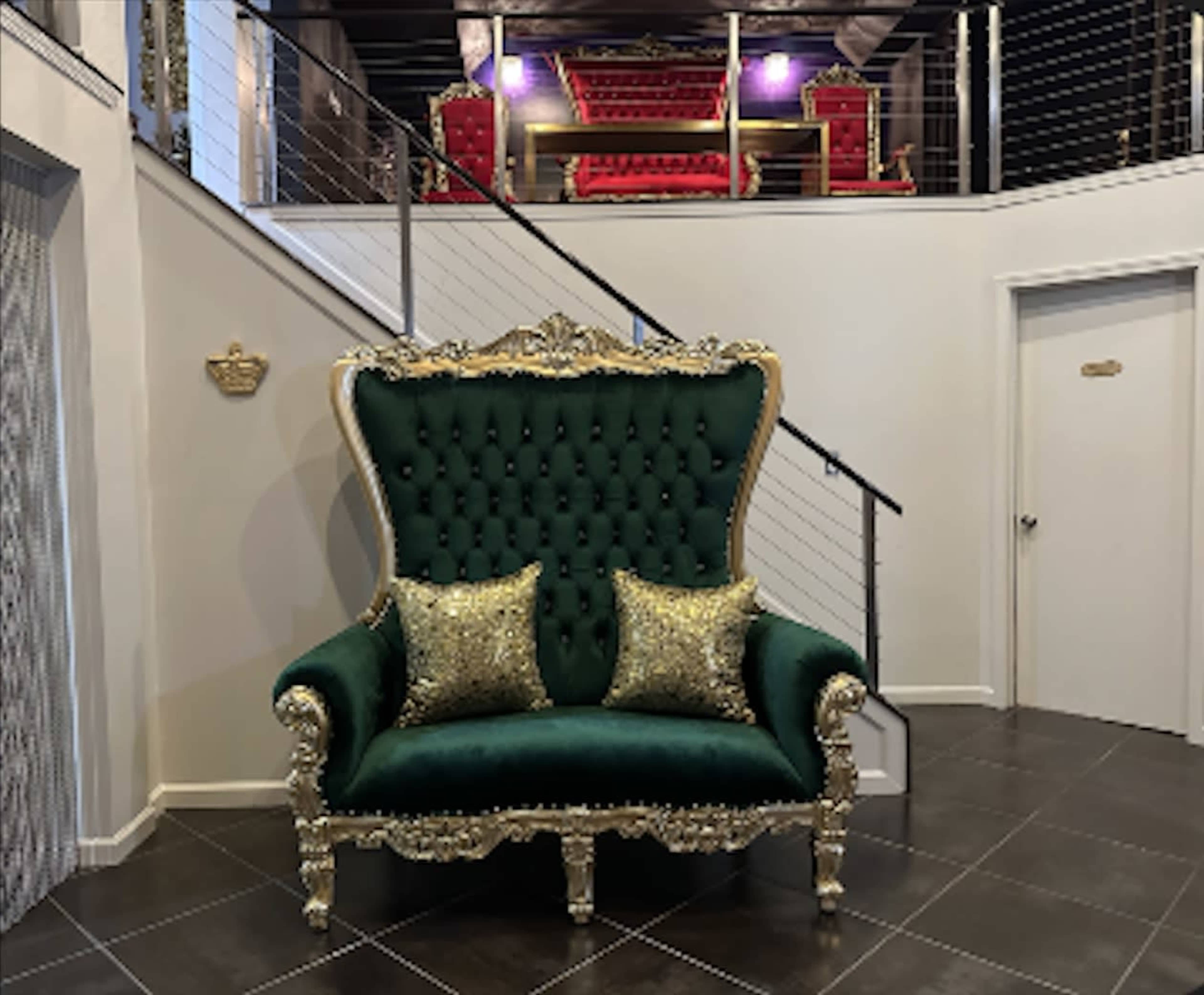 A large, ornate green velvet chair with gold accents and two decorative pillows sits in front of a staircase leading to a second floor with red upholstered furniture.