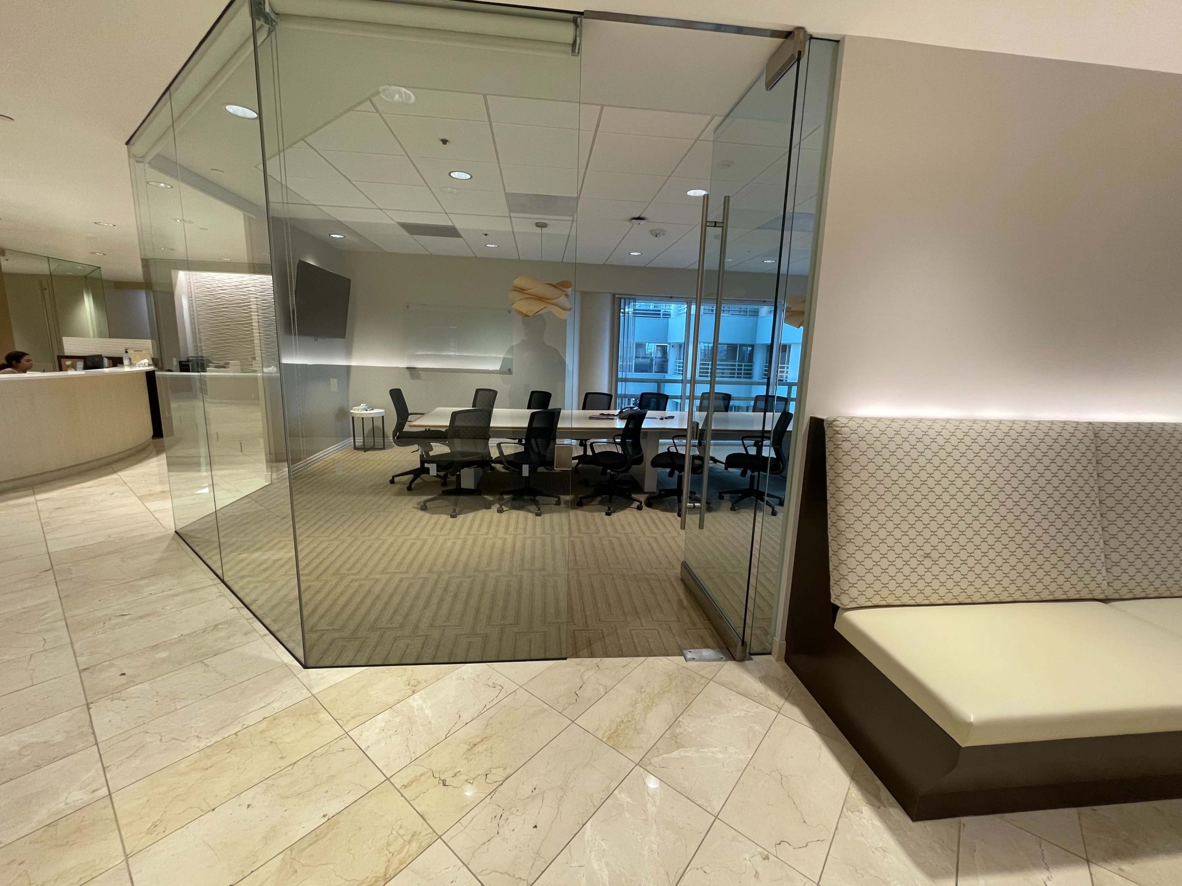 The image shows a modern office space with a glass-walled conference room containing several chairs and a large table, adjacent to a reception area.