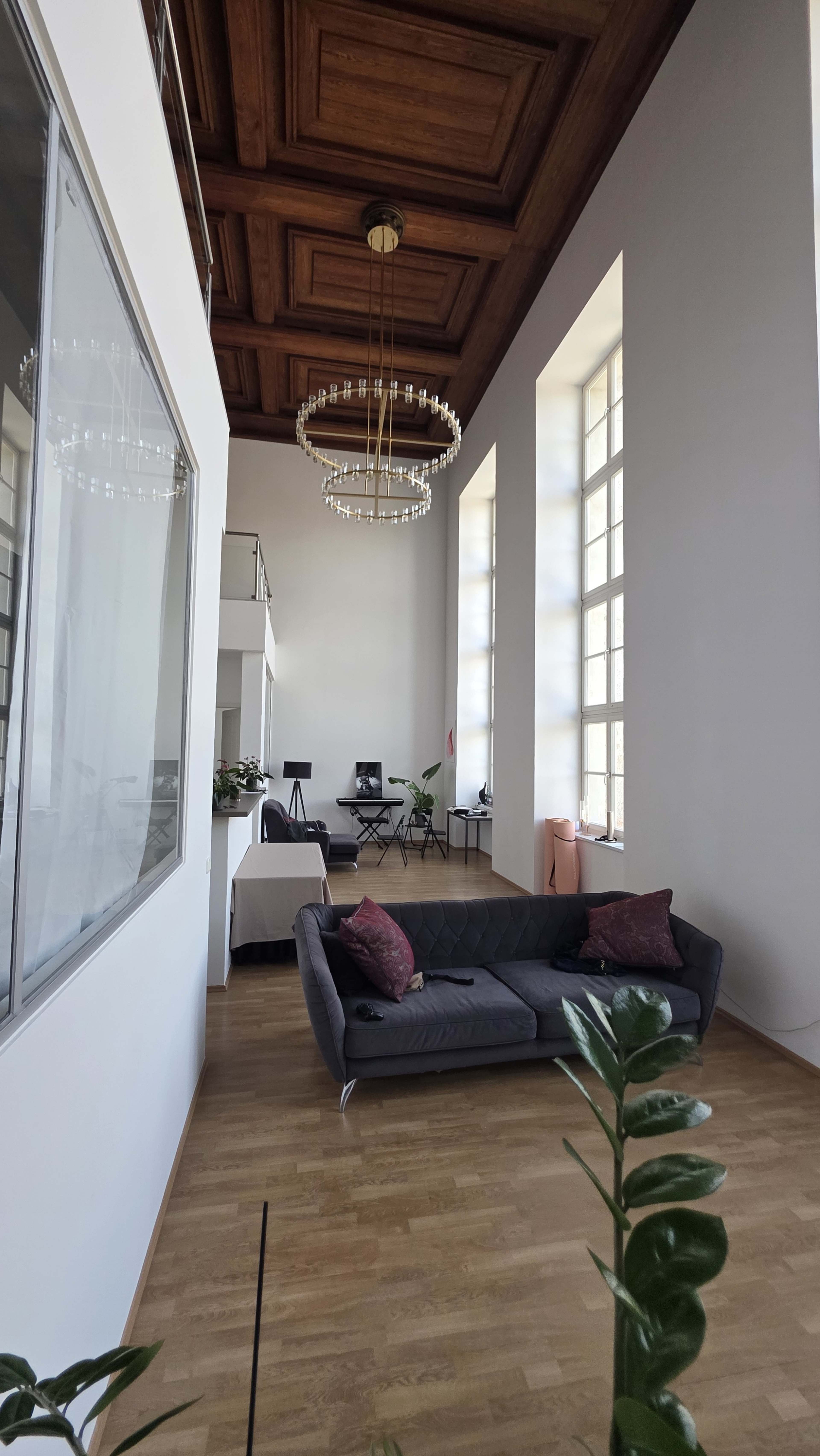 The image shows a spacious interior with high ceilings, featuring a dark couch, large windows, and a chandelier.