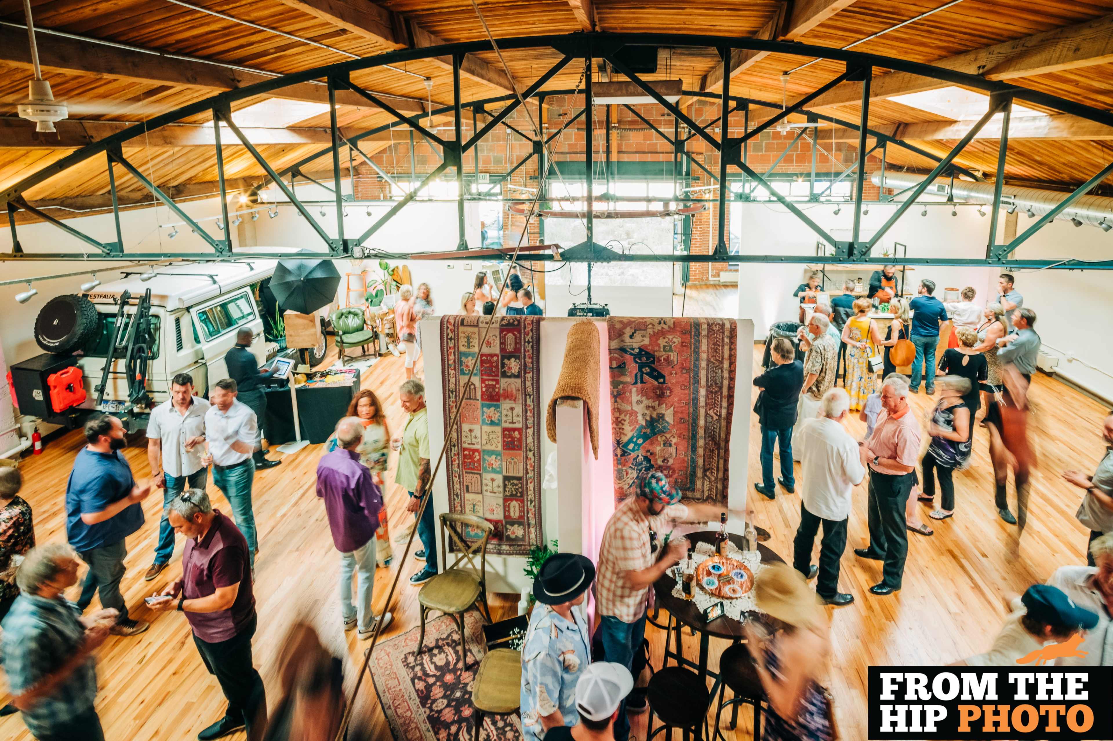 A lively gathering takes place in a spacious, brightly lit venue with attendees mingling around tables and displays.