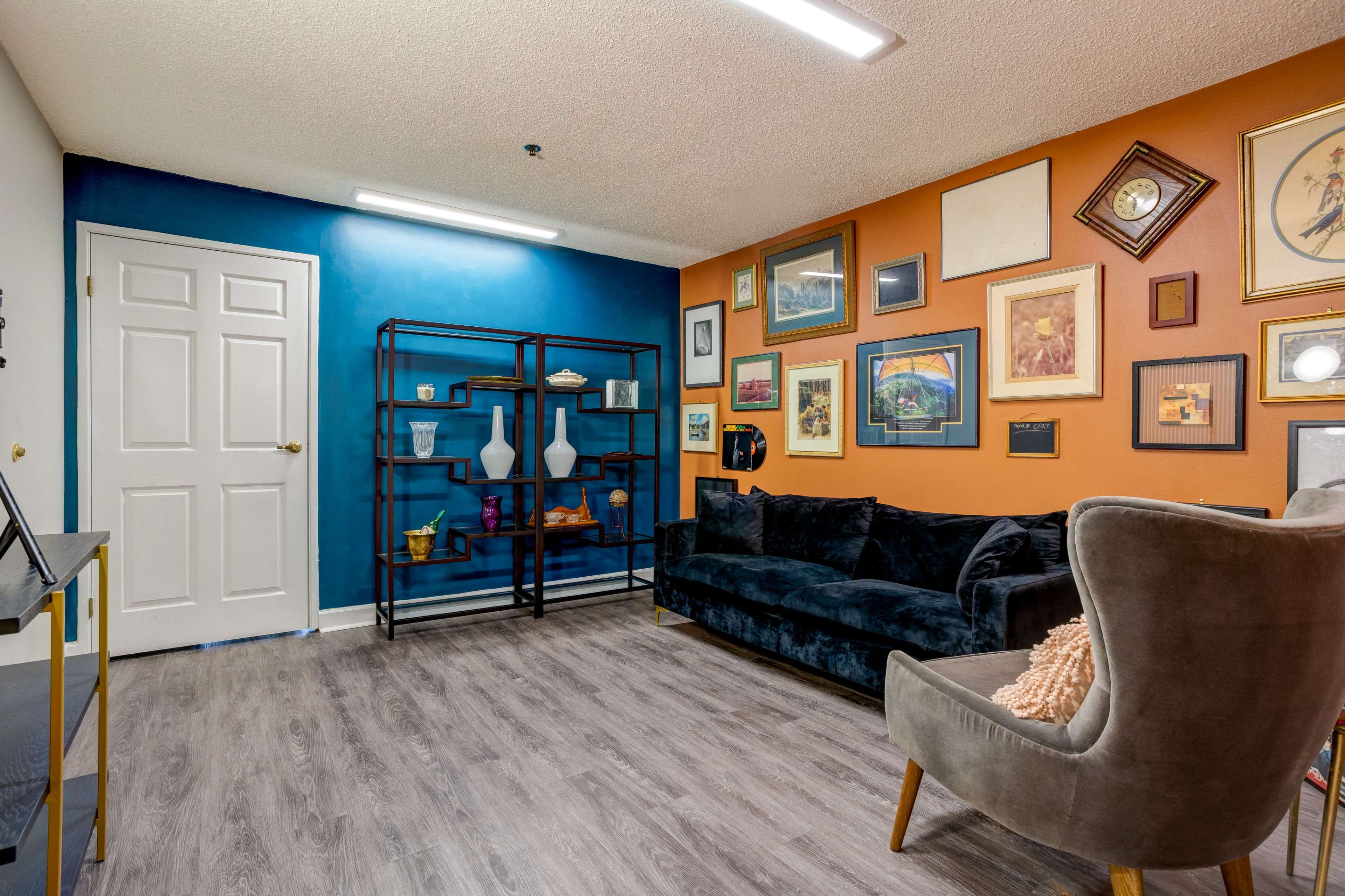 The room features a black couch and a grey armchair, with an assortment of framed artwork displayed on the orange and blue walls.