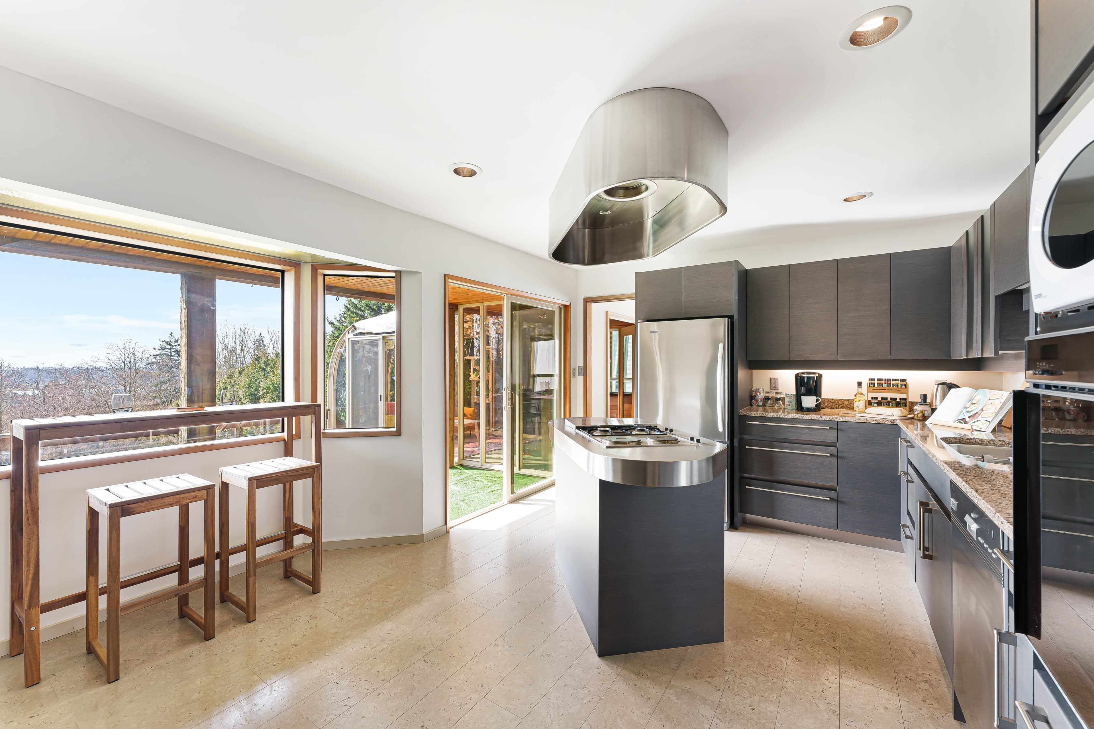 A modern kitchen features sleek cabinets, a central island with a stovetop, and large windows that allow natural light to fill the space.