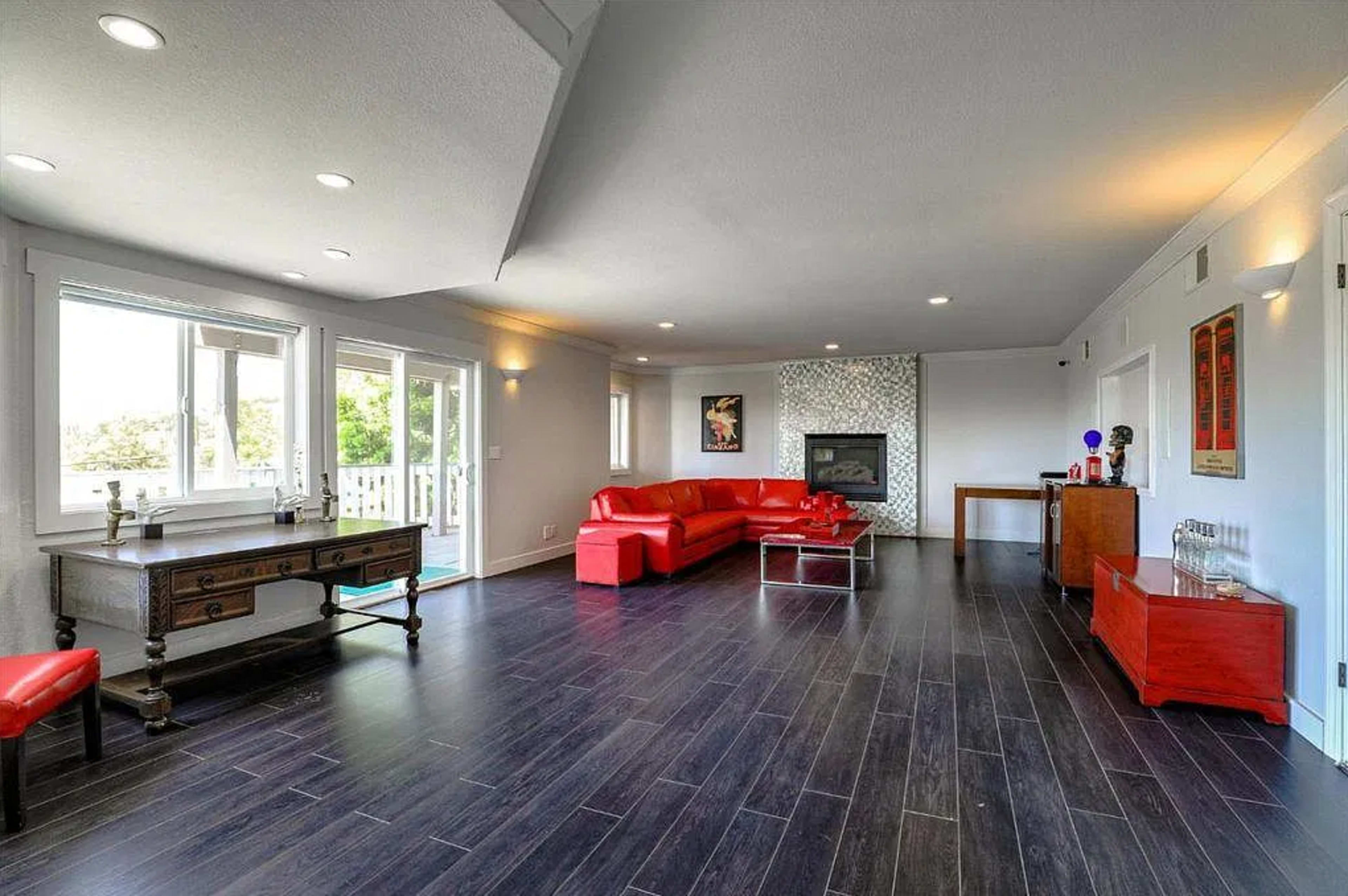 The image shows a modern living room featuring a red sectional sofa, a fireplace, and dark wooden flooring, with large windows allowing natural light to enter.