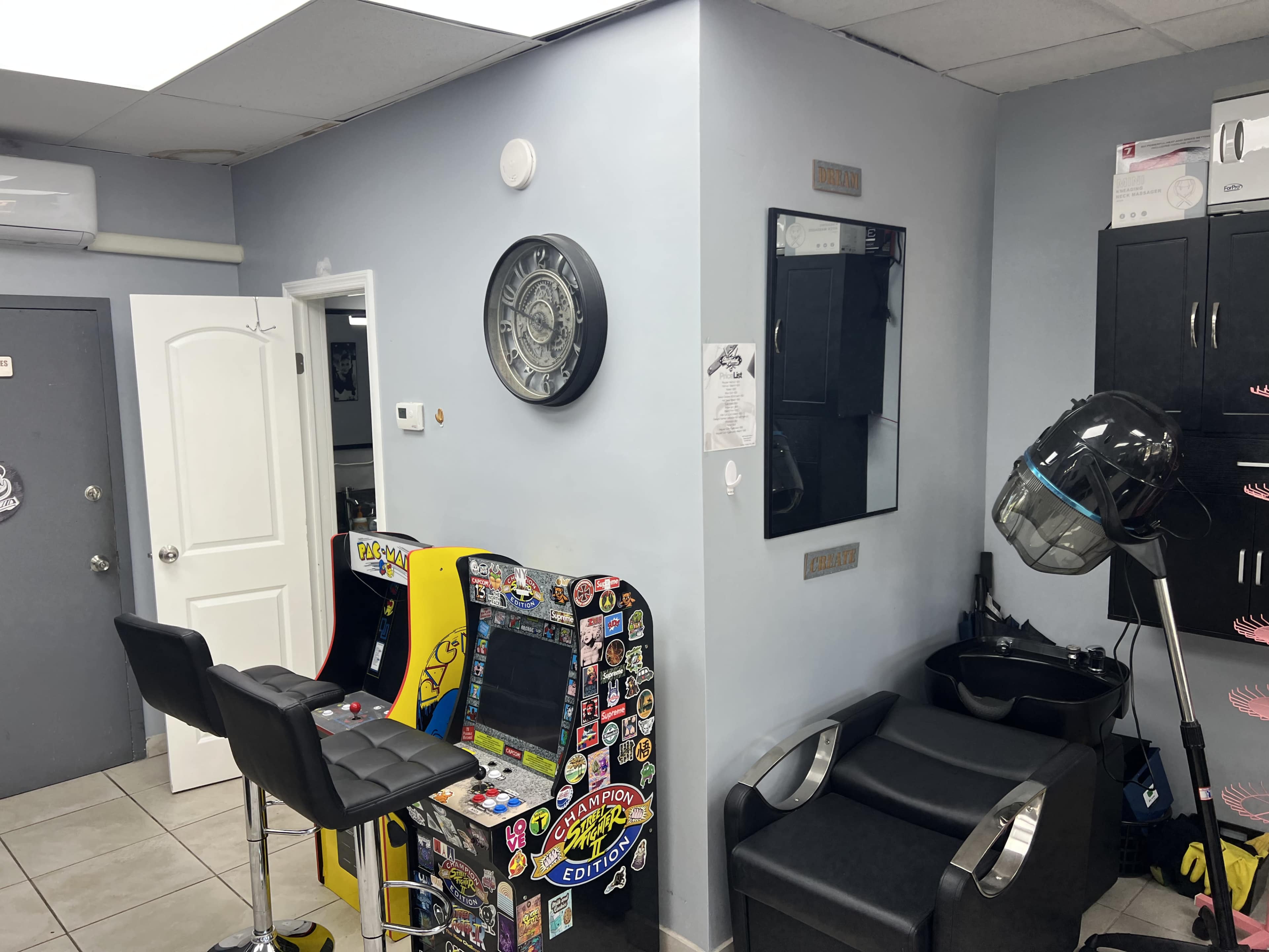 The image shows a barbershop interior featuring two black chairs, a vintage arcade game machine covered in stickers, and a wall clock.