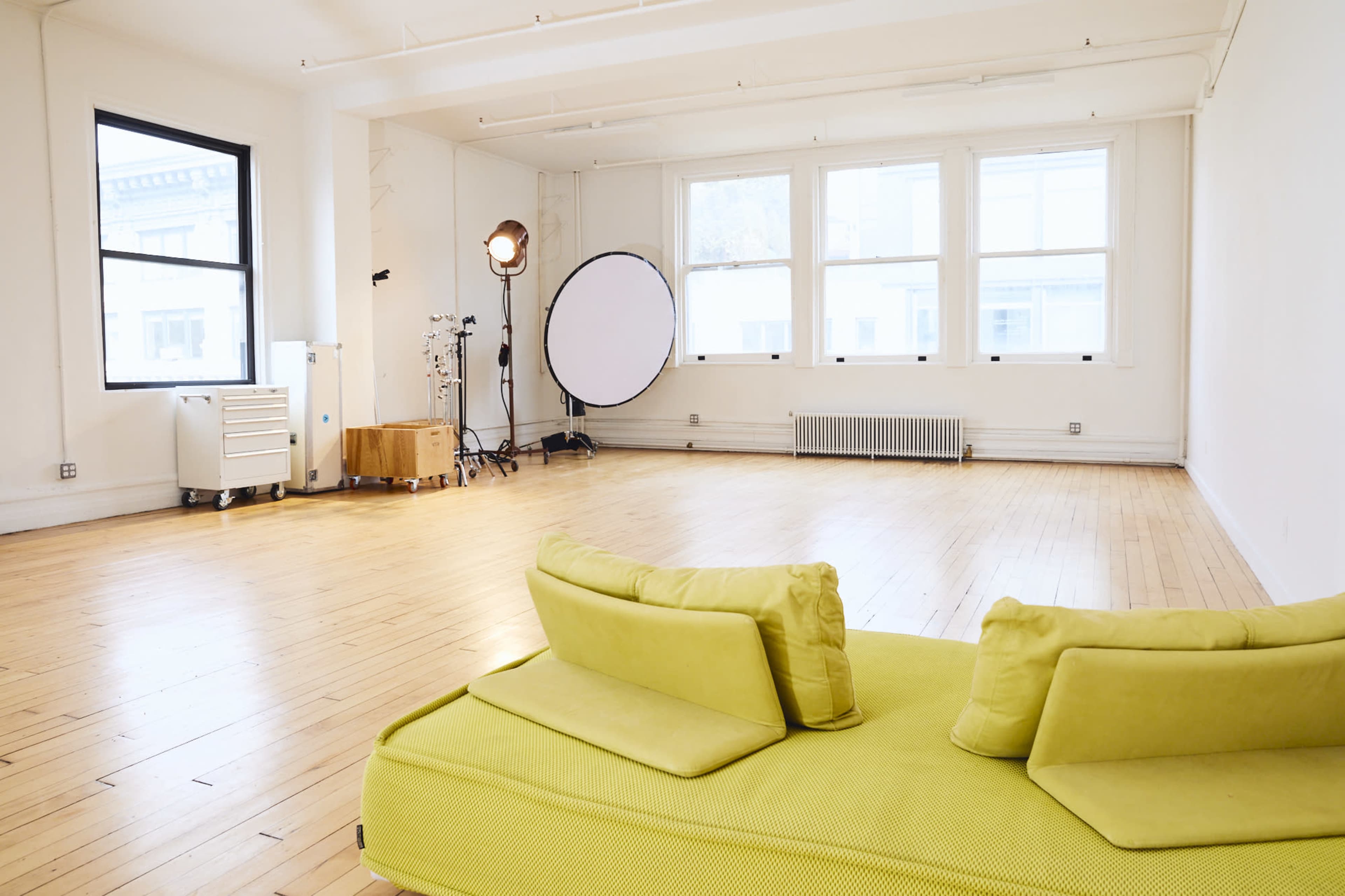 The image shows a bright, empty studio space with wooden floors, large windows, a green couch, and lighting equipment in the corner.