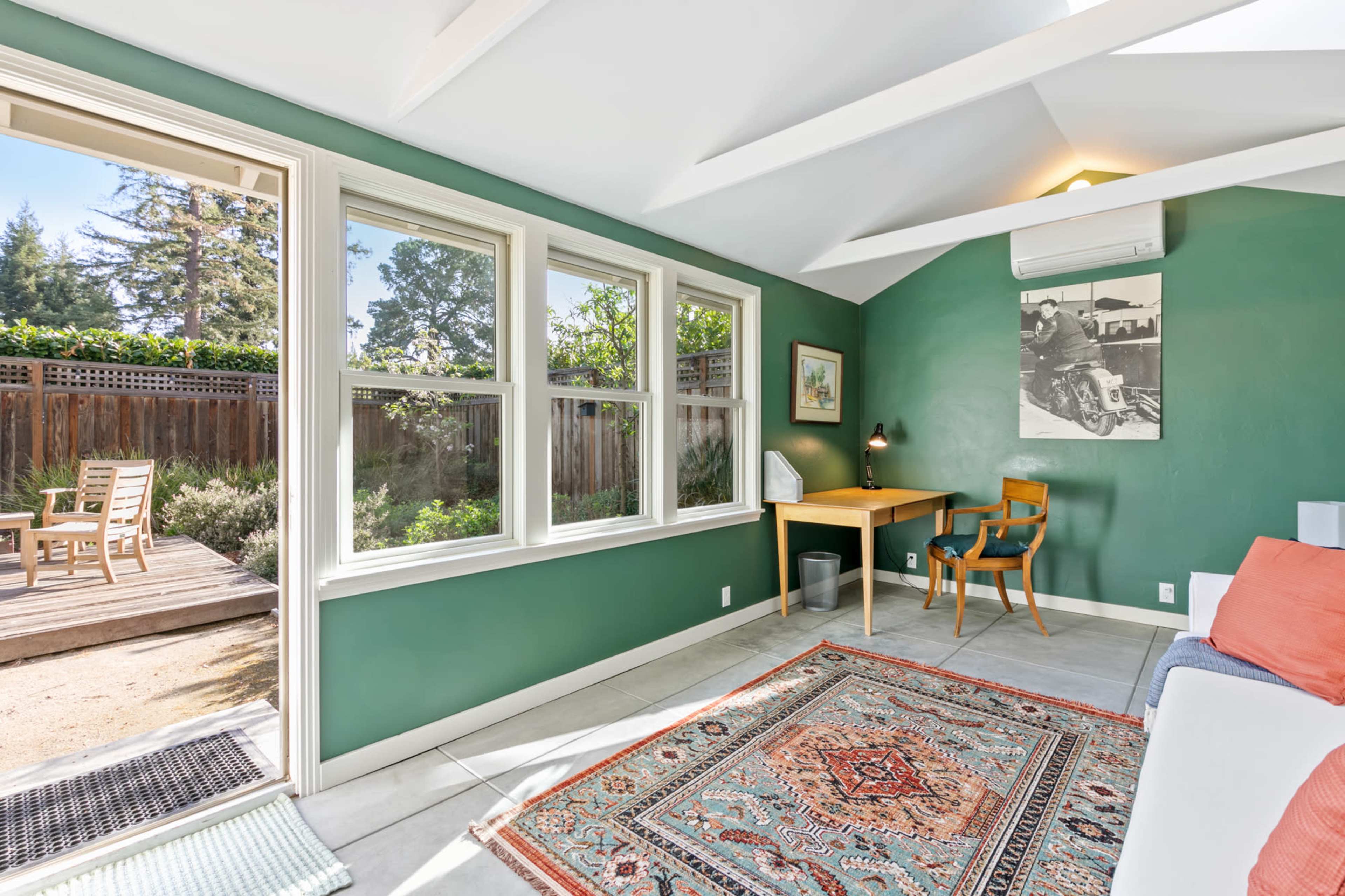 A bright, green-walled living space with large windows, a desk and chair, and a patio area visible through an open door.