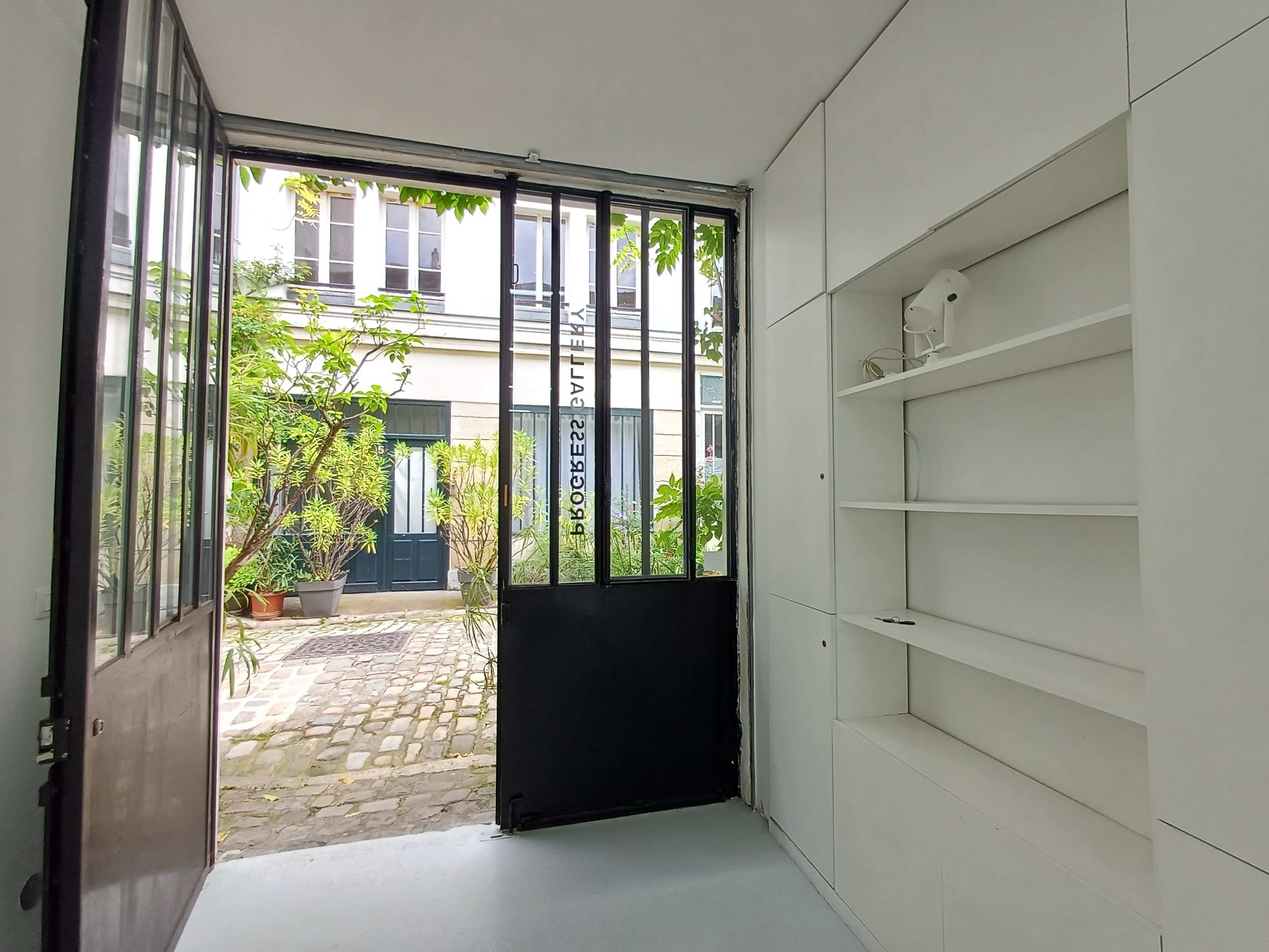 A doorway leads from a minimalist interior to a cobblestone courtyard adorned with potted plants and green foliage.