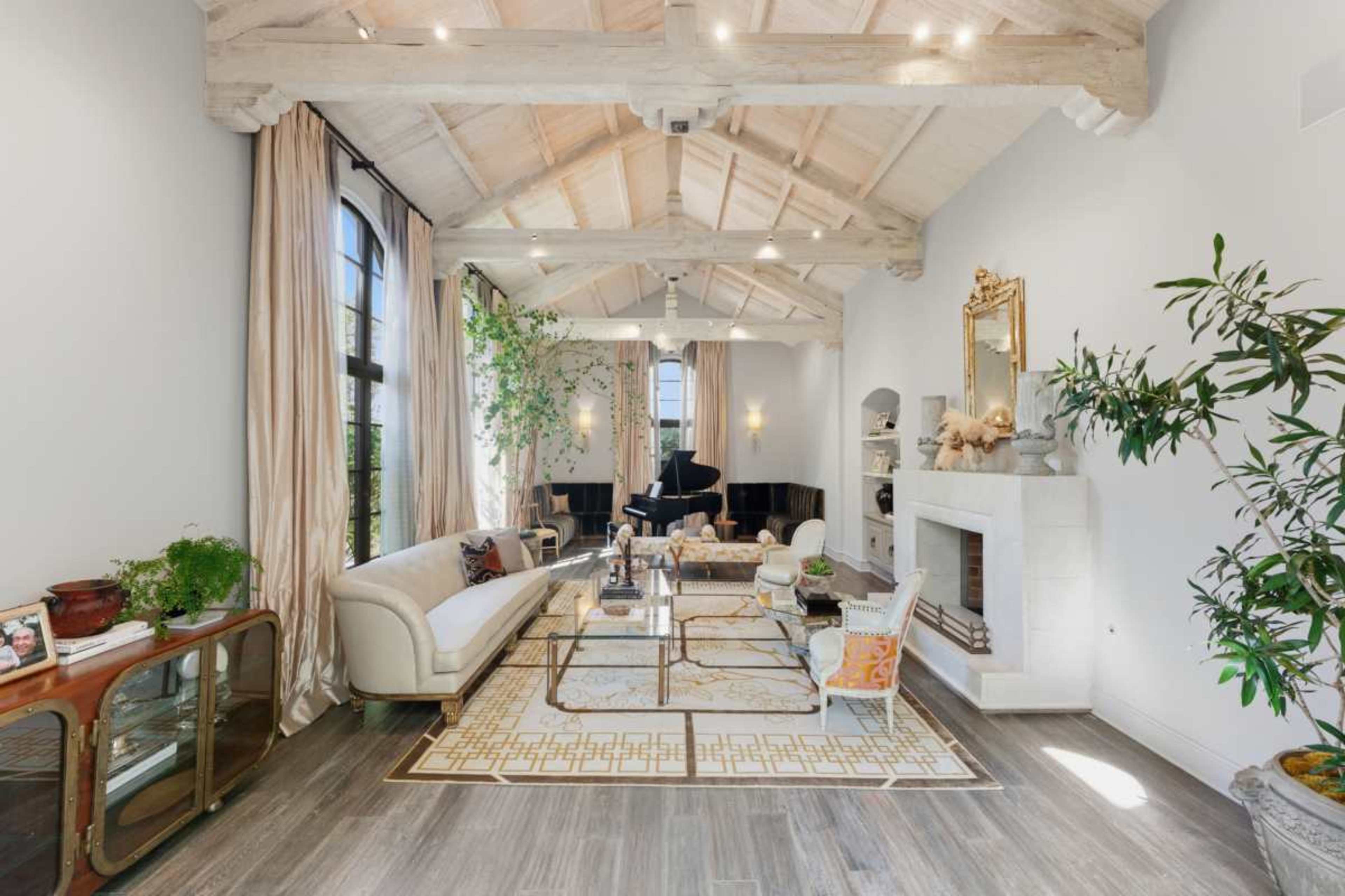 The image depicts a spacious living room featuring high ceilings with wooden beams, a grand piano, and large windows, with a mix of indoor plants and elegant furnishings.