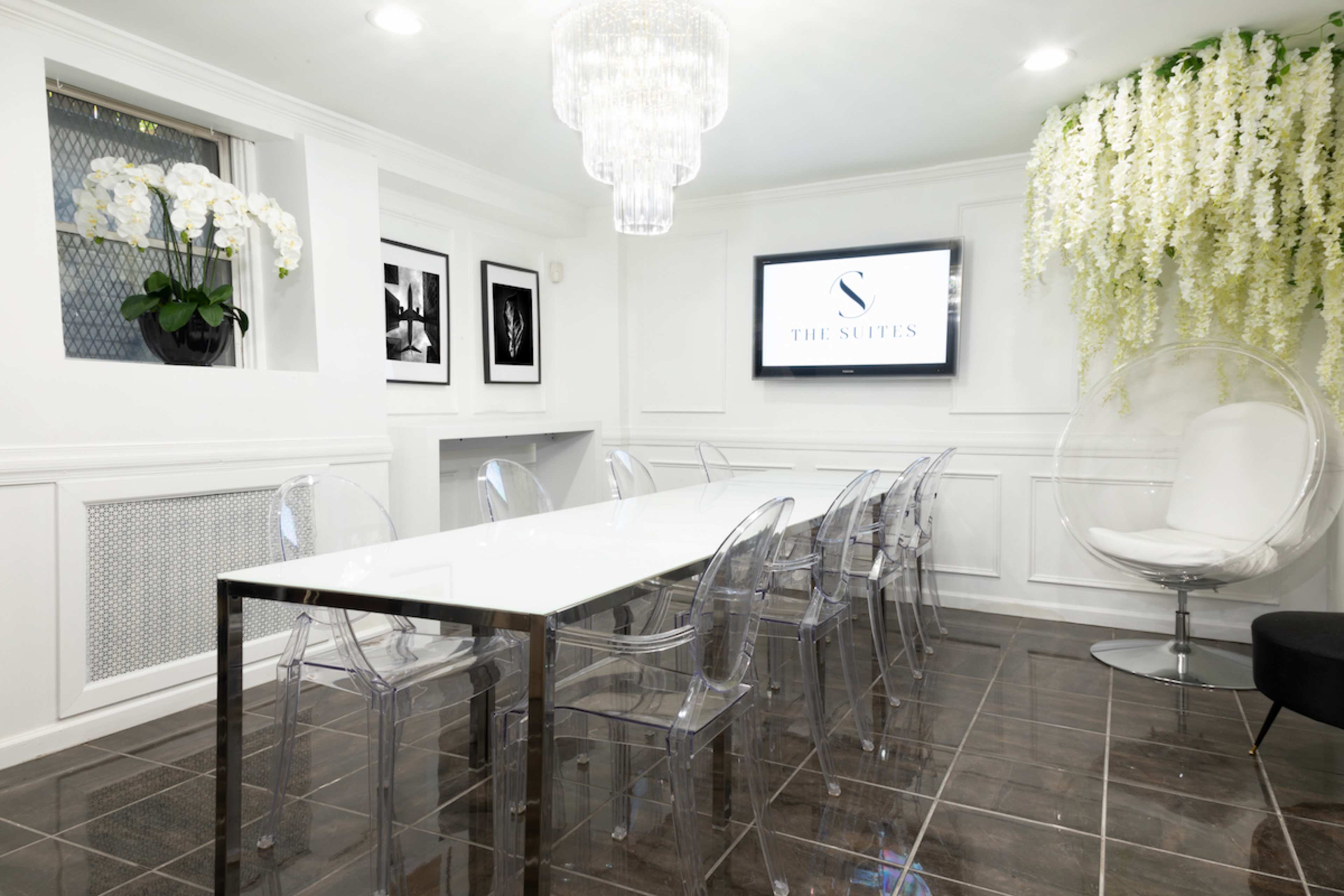 A modern conference room with a glass table surrounded by clear acrylic chairs, a large TV on the wall, and decorative plants hanging from the ceiling.