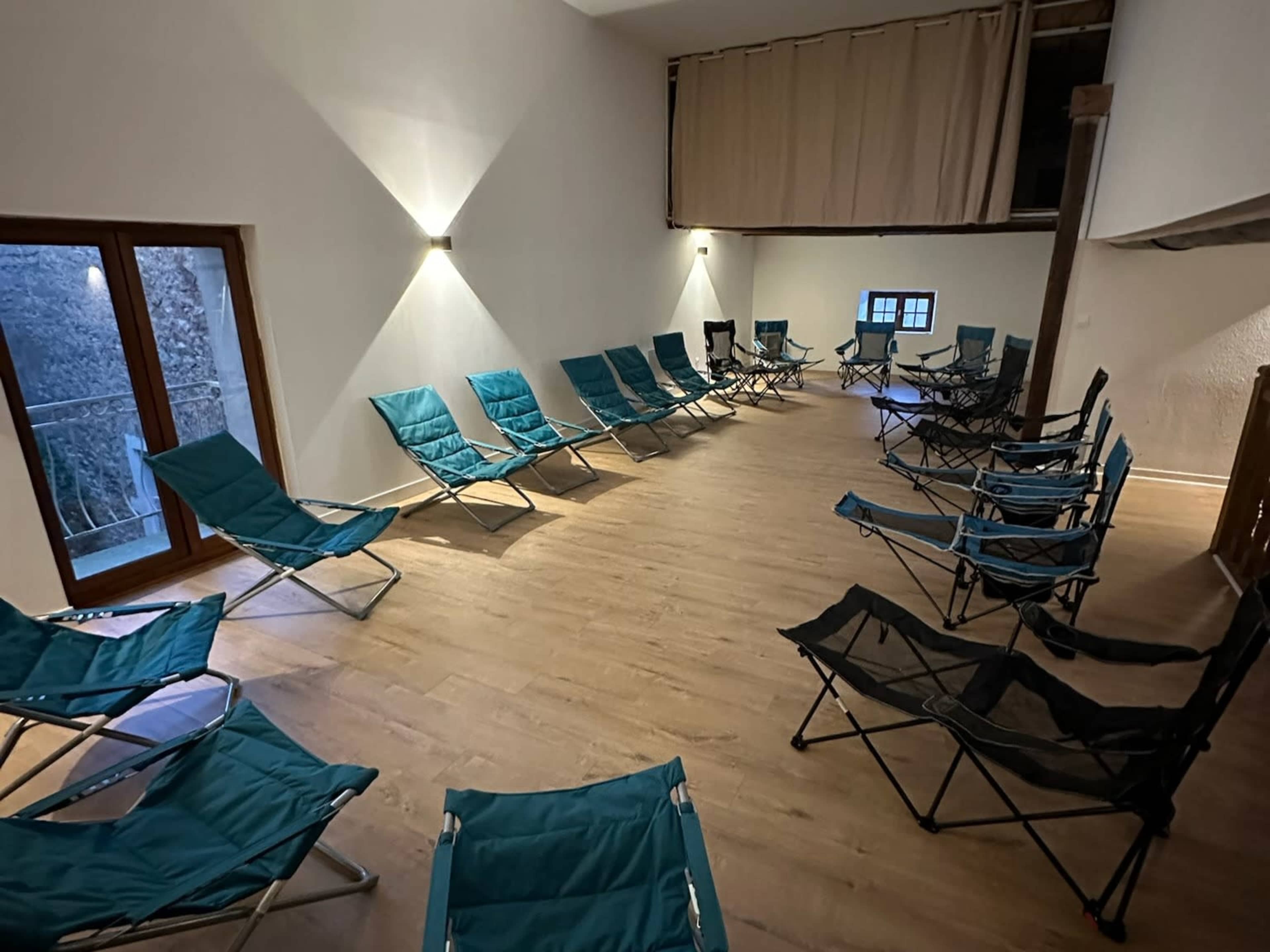 A spacious room features a circular arrangement of blue foldable chairs and large windows allowing natural light.