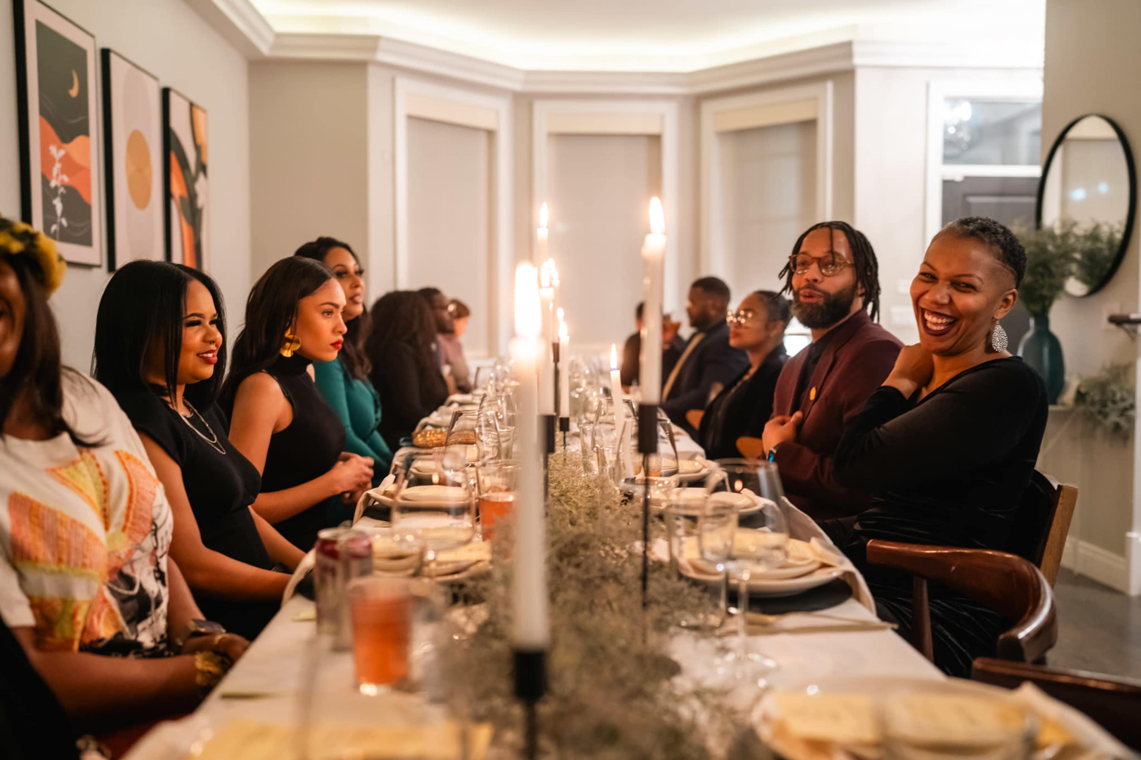 A group of elegantly dressed people gather around a long dining table adorned with candles and table settings in a well-lit room.
