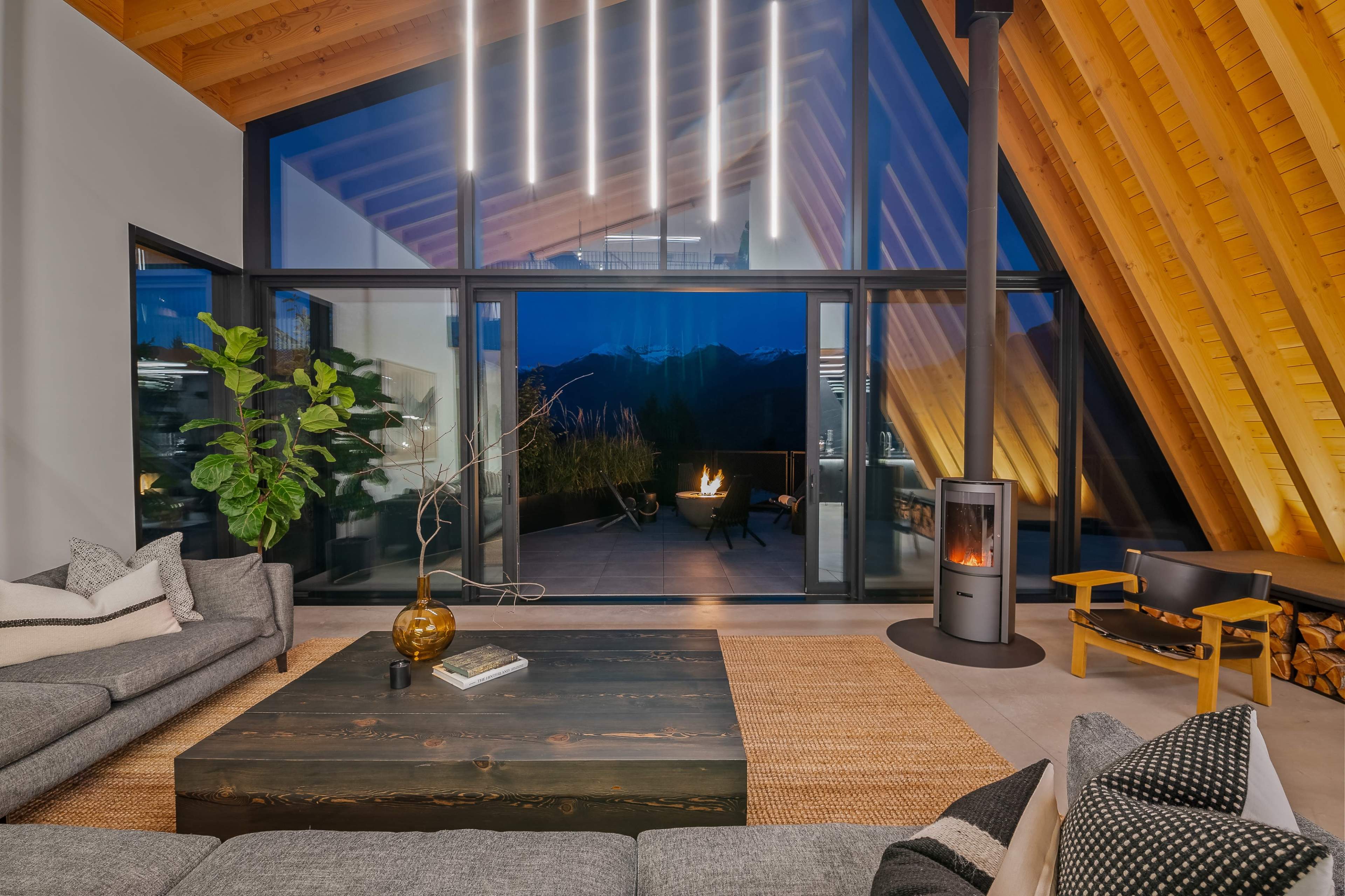 The image shows a modern living space with large windows overlooking a mountain view, featuring a cozy seating area, a central wooden coffee table, and a wood-burning stove.