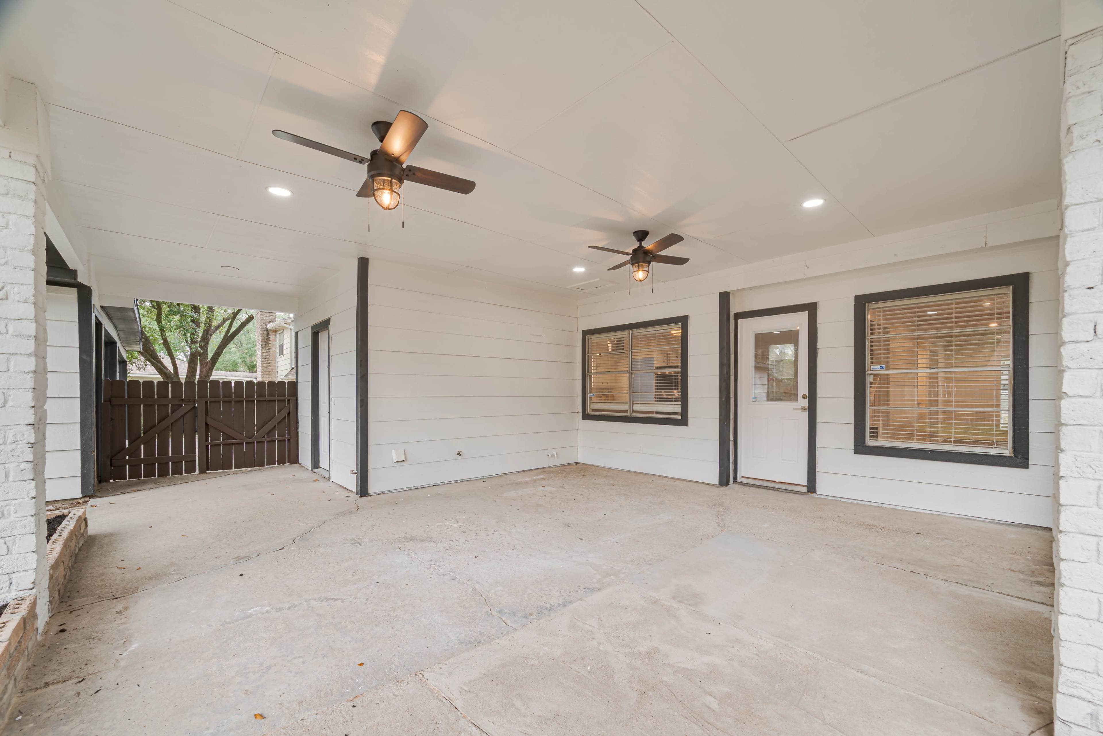 The image shows a spacious covered patio with two ceiling fans, light fixtures, and a wooden fence in the background.