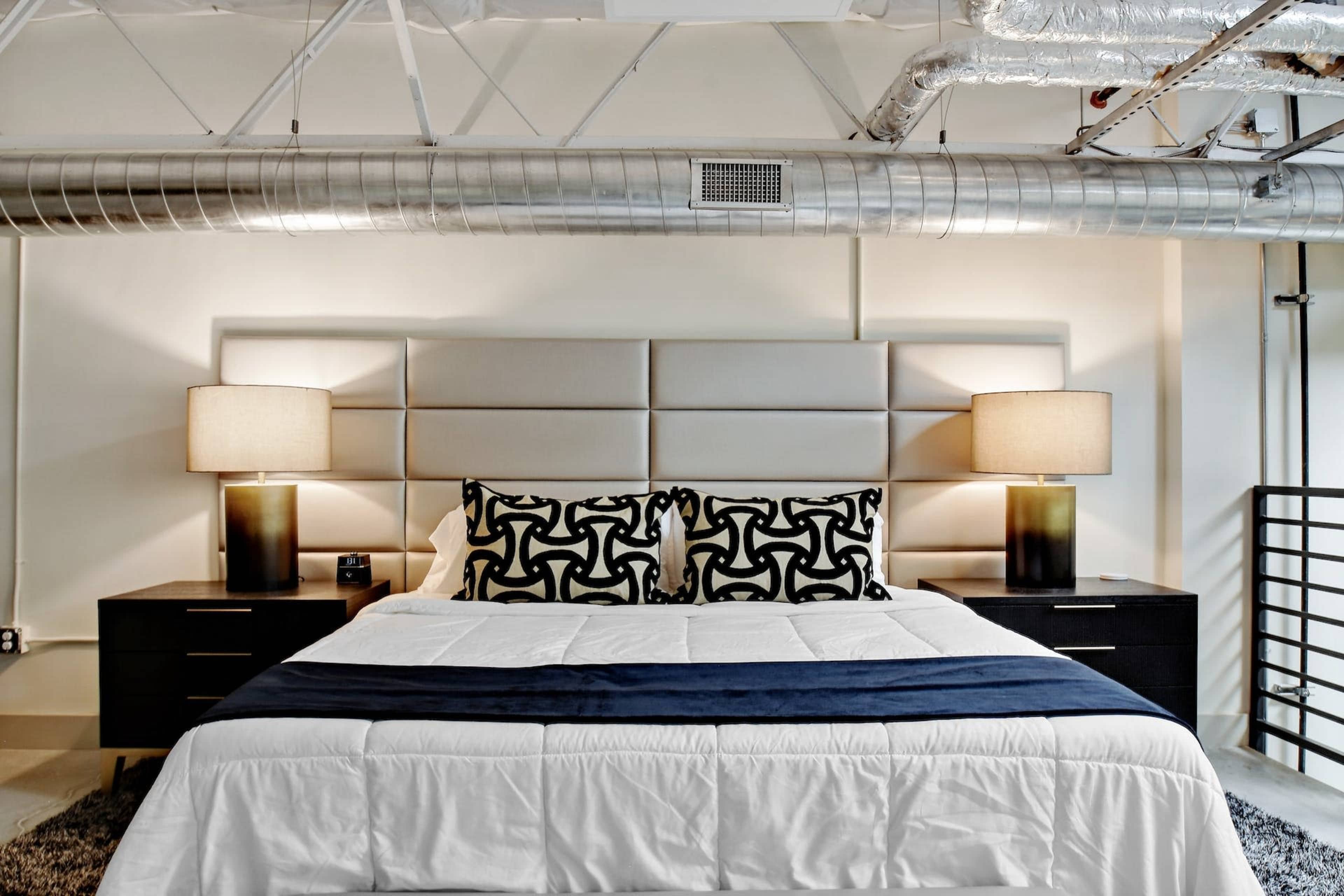 A modern bedroom features a large bed with a white comforter, flanked by two lamps and dark wood nightstands.