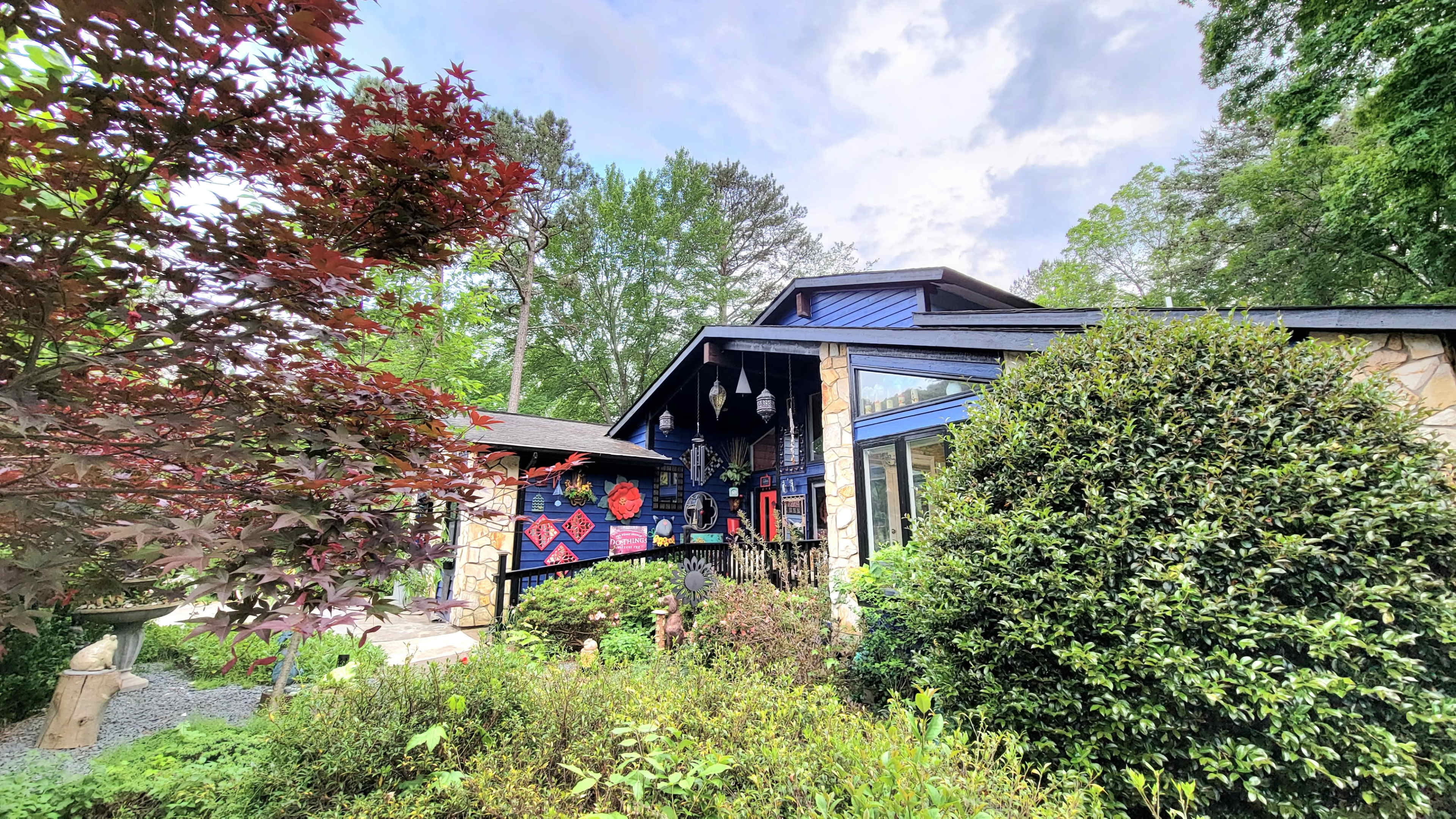 A blue house with stone accents and decorative items is surrounded by lush greenery and colorful plants.