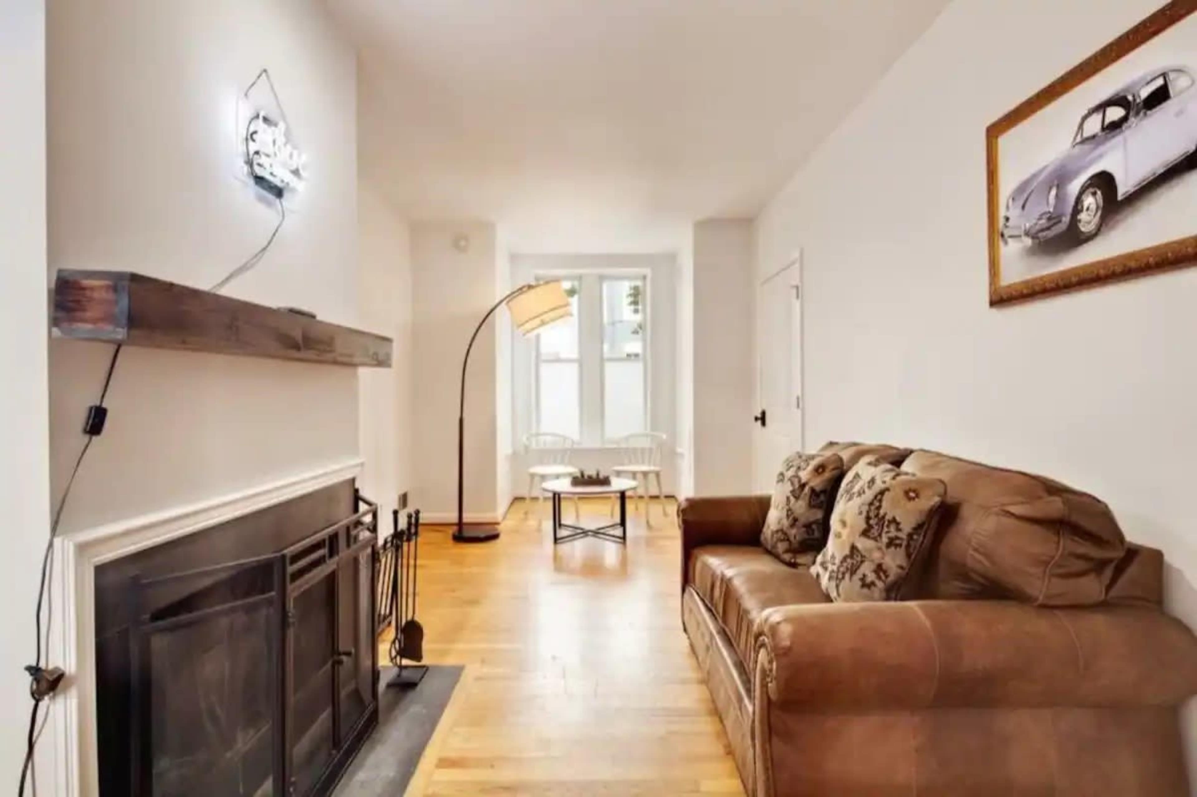 A living room with a brown leather sofa, a fireplace, a floor lamp, and a framed picture of a vintage car on the wall.