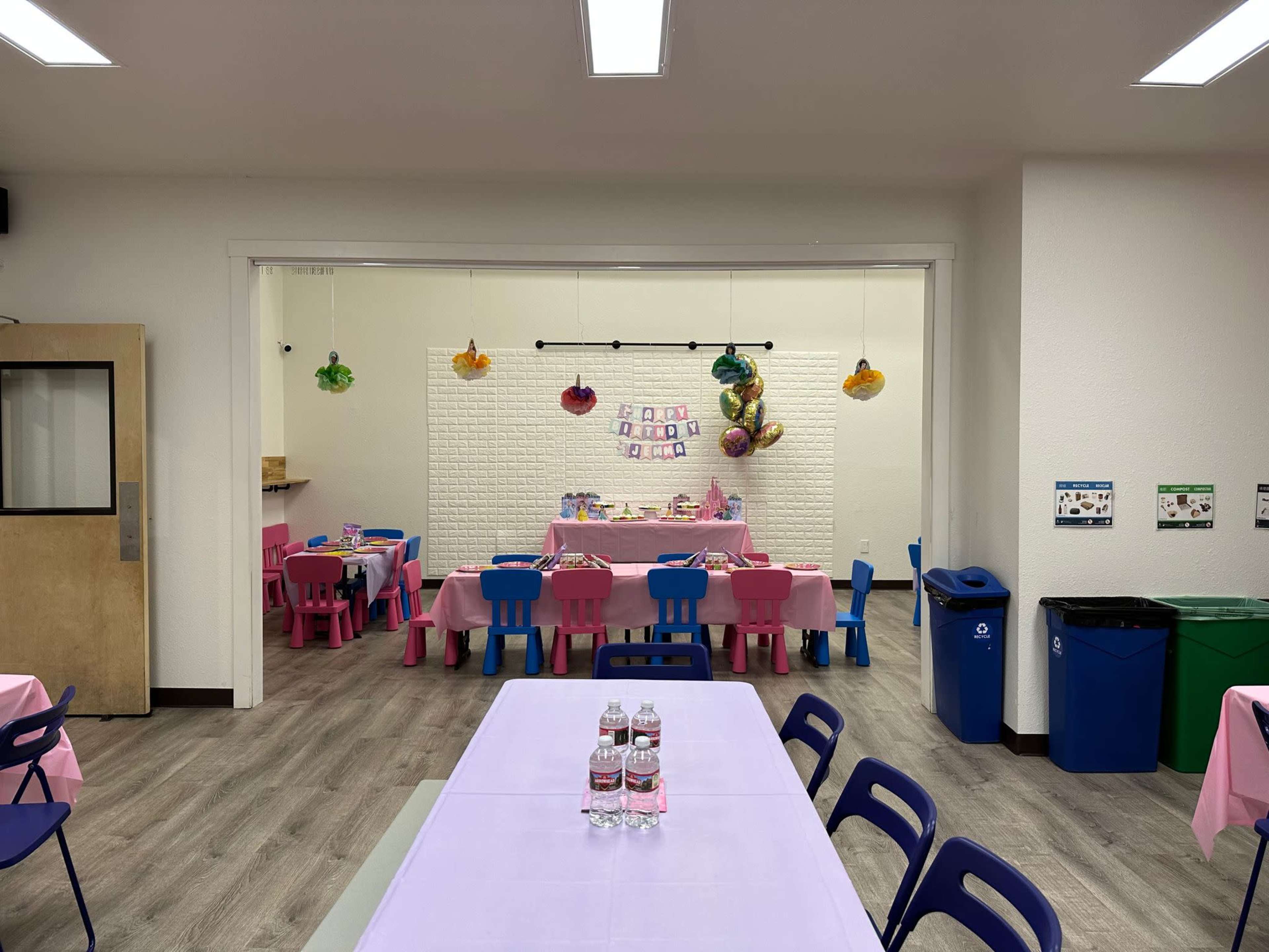 A brightly decorated room with pink and blue tables set for a party, featuring colorful paper lanterns and a dessert table in the background.