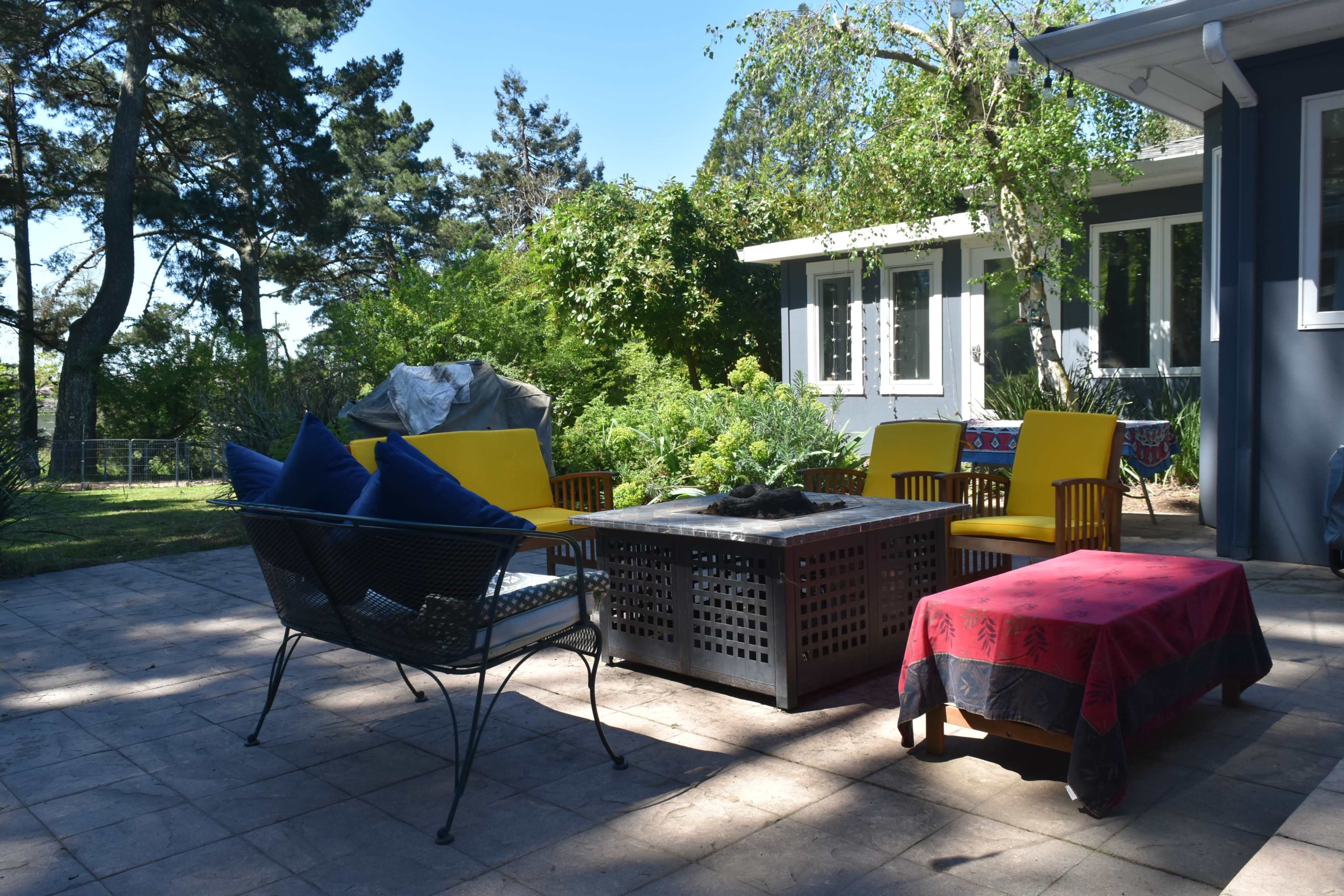 A patio area features a table with a fire pit surrounded by yellow chairs and a red bench, with greenery and a house in the background.