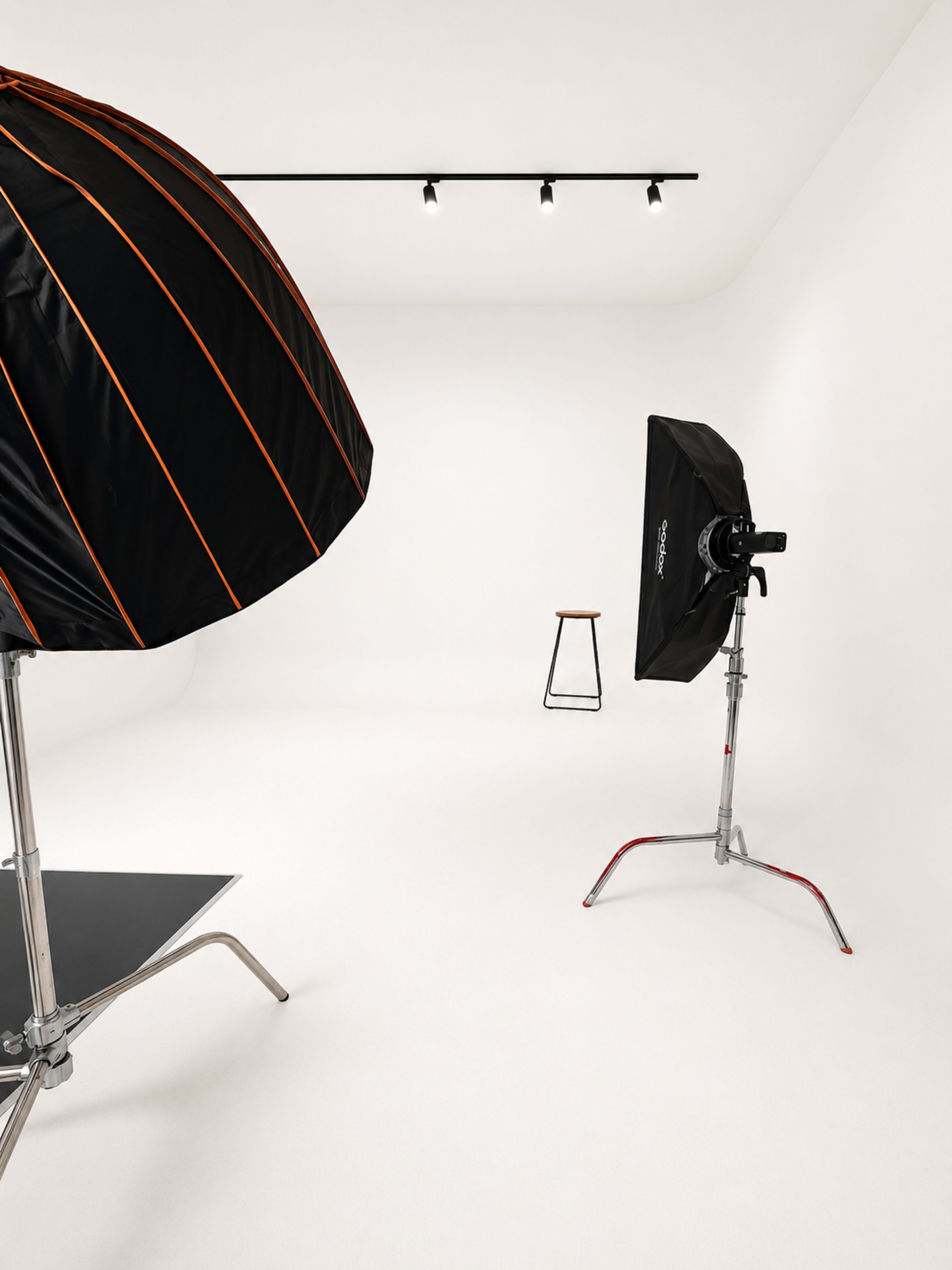 The image shows a minimalist photography studio featuring a black and orange softbox light, a black umbrella reflector, and a single black stool against a white background.
