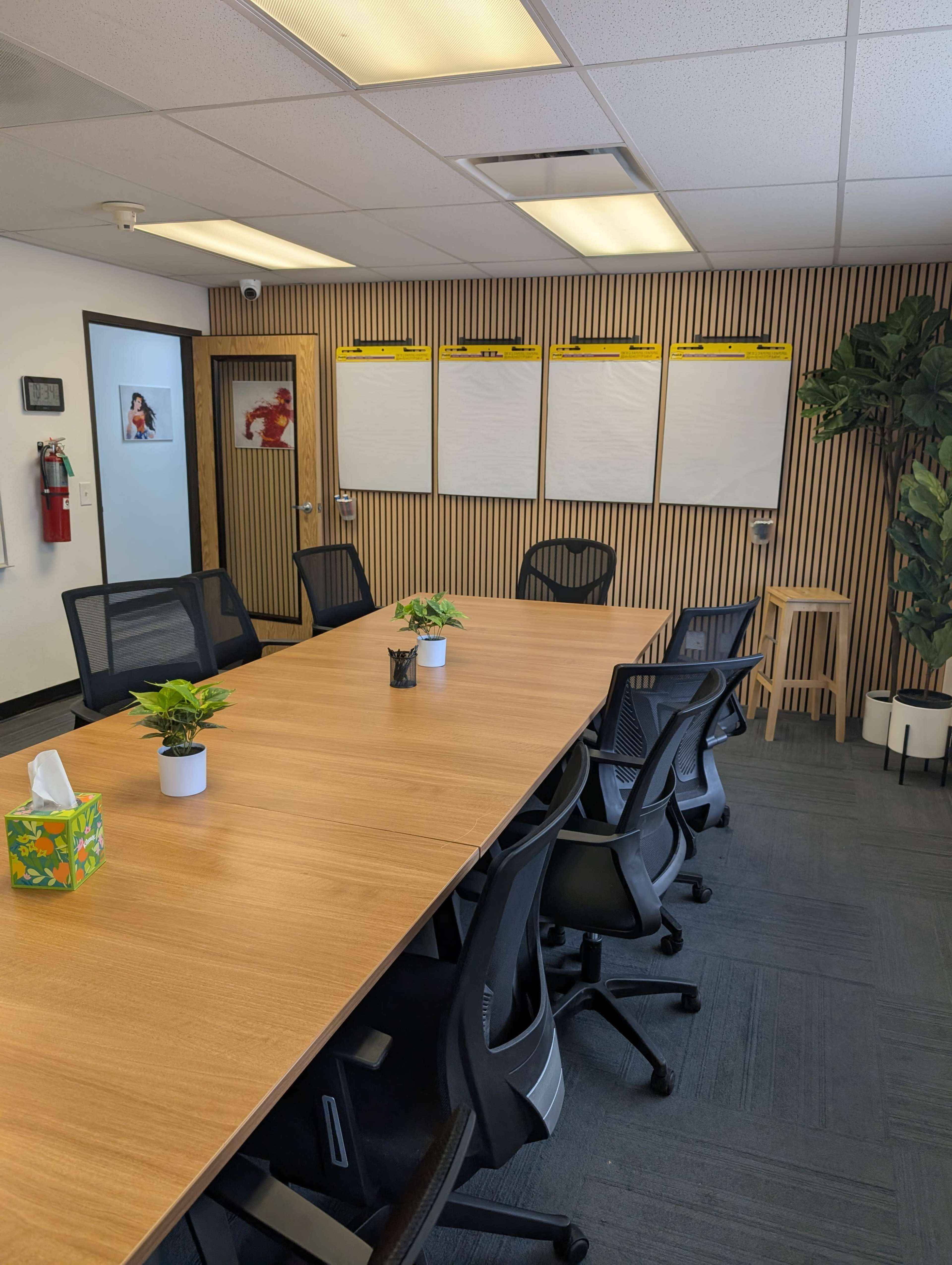 A conference room features a large wooden table surrounded by black chairs, with a plant in each corner and flip charts mounted on the wall.