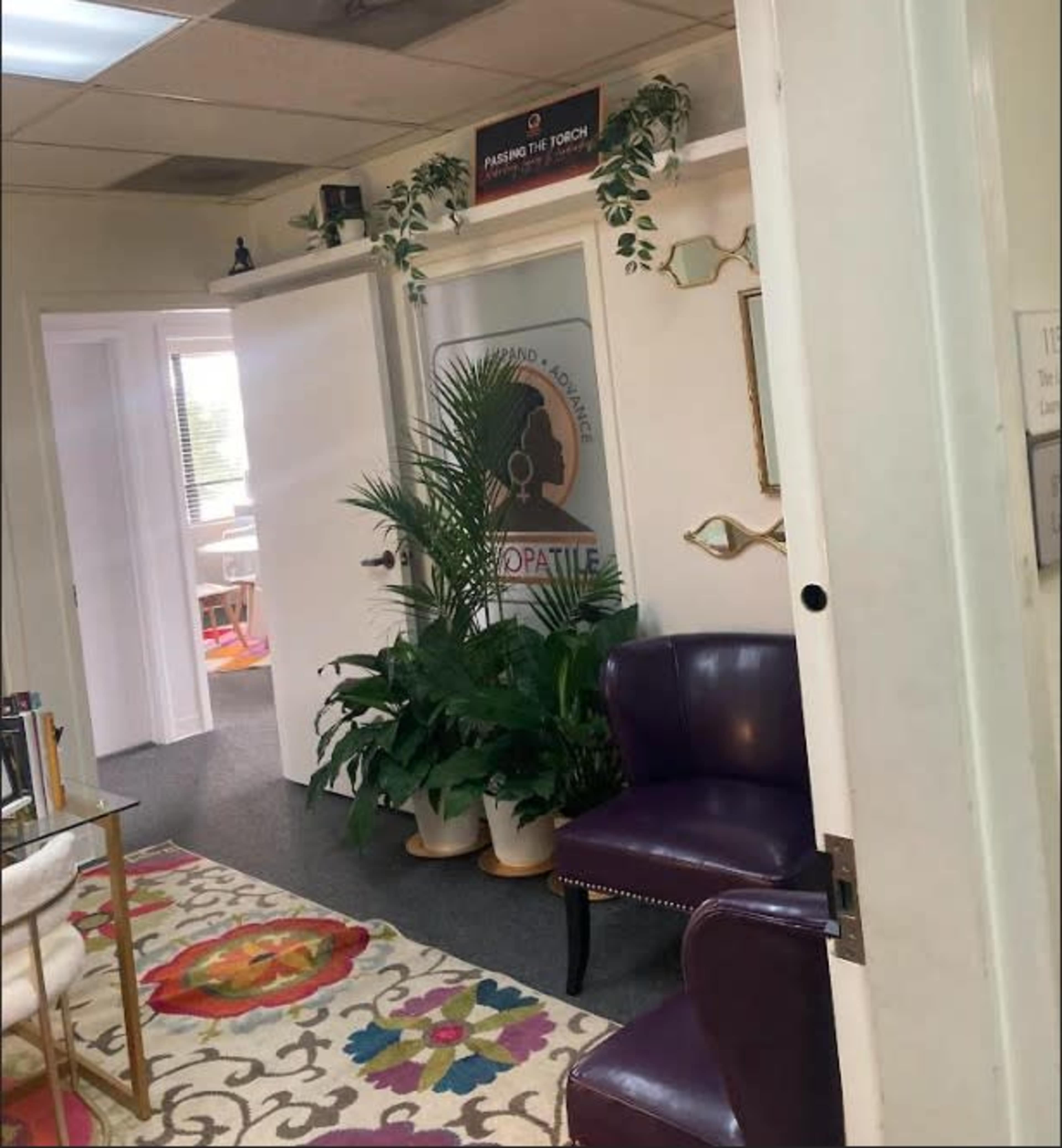 The image shows an office entrance with a sign, a purple armchair, potted plants, and a colorful patterned rug.
